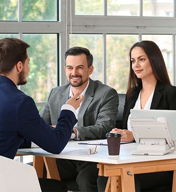 Three colleagues in a bright office discussing work at a table with a laptop and coffee cup