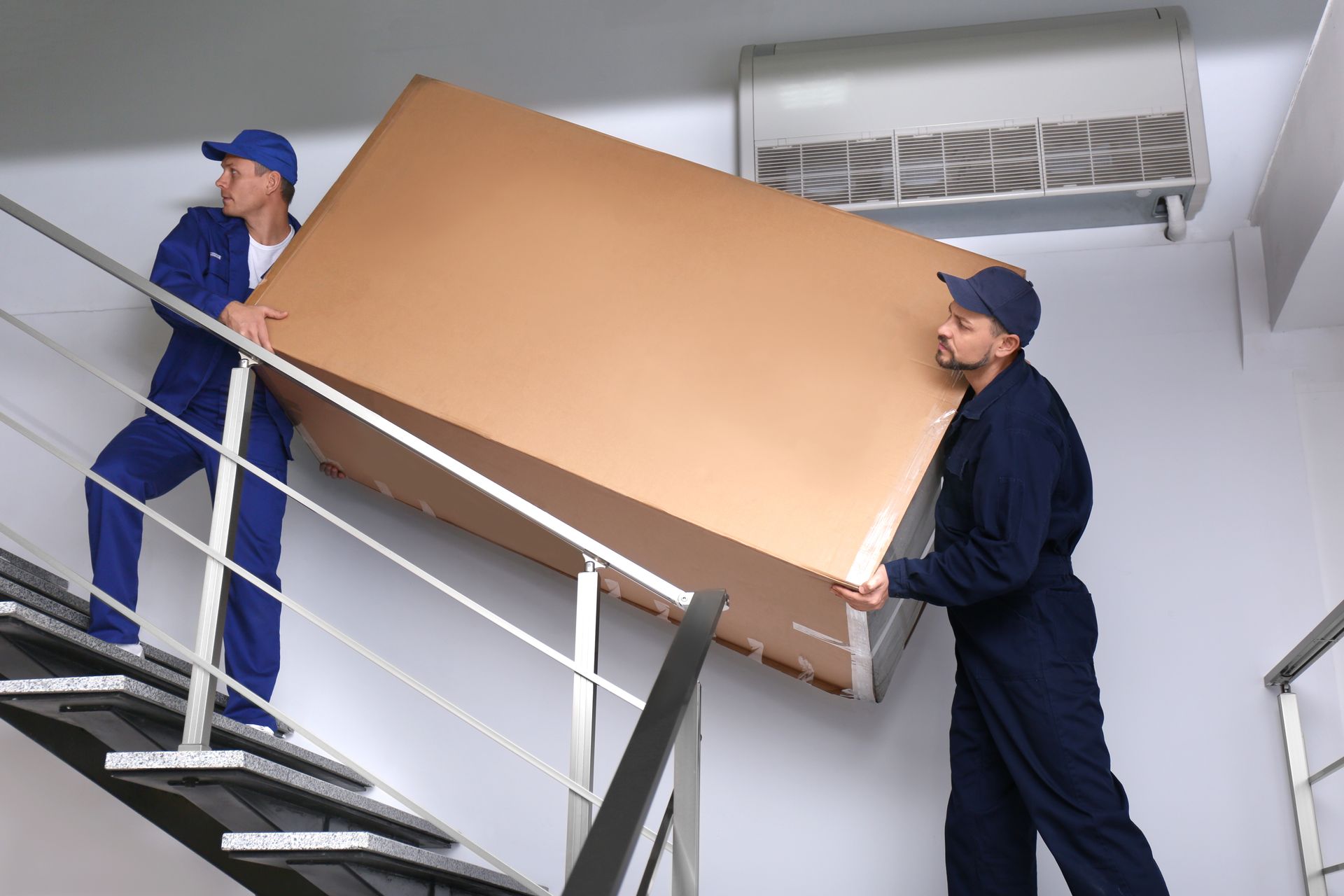 Two men are carrying a large cardboard box up a set of stairs.