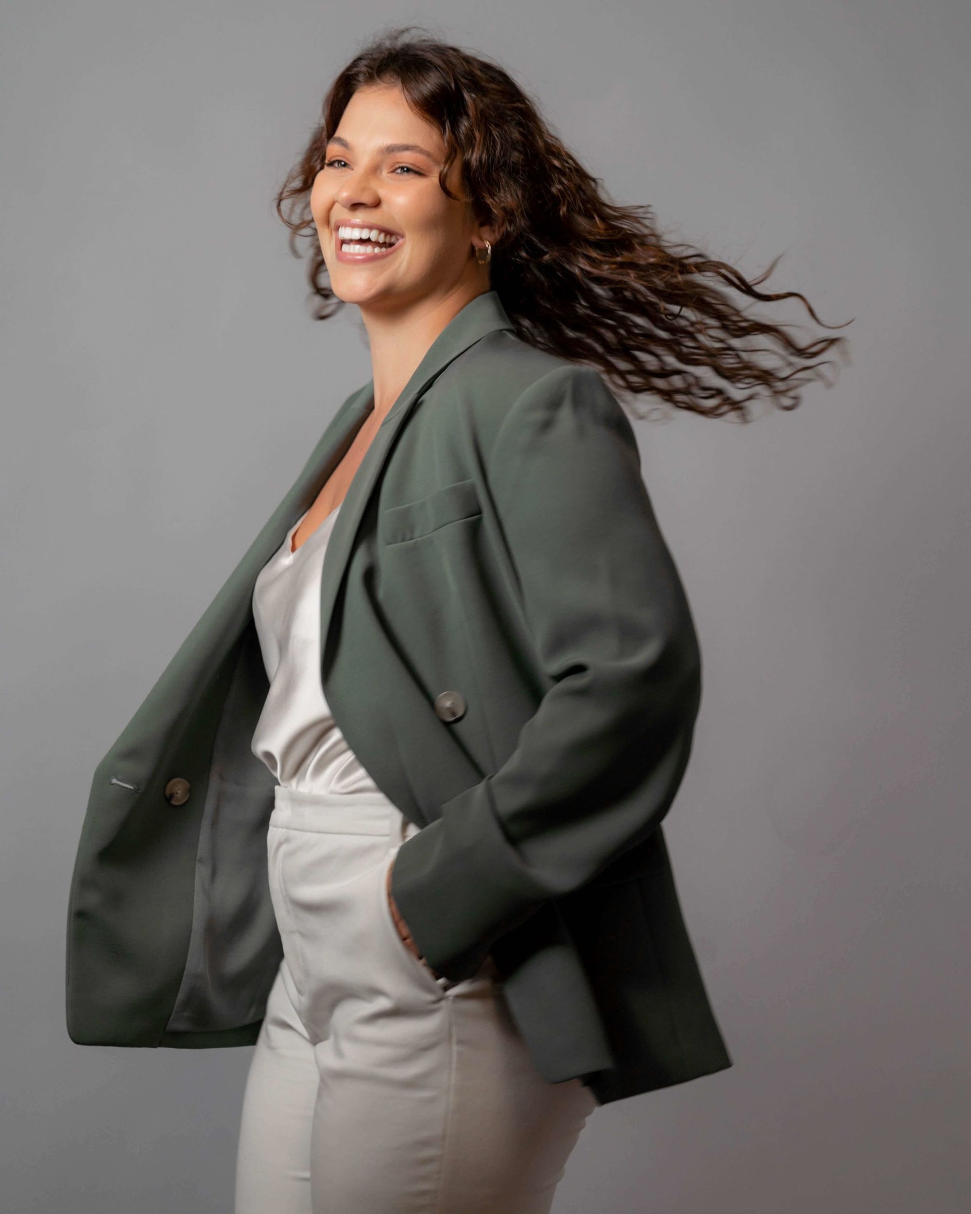 Woman with curly hair smiles, wearing a green blazer and white outfit, against a gray backdrop.