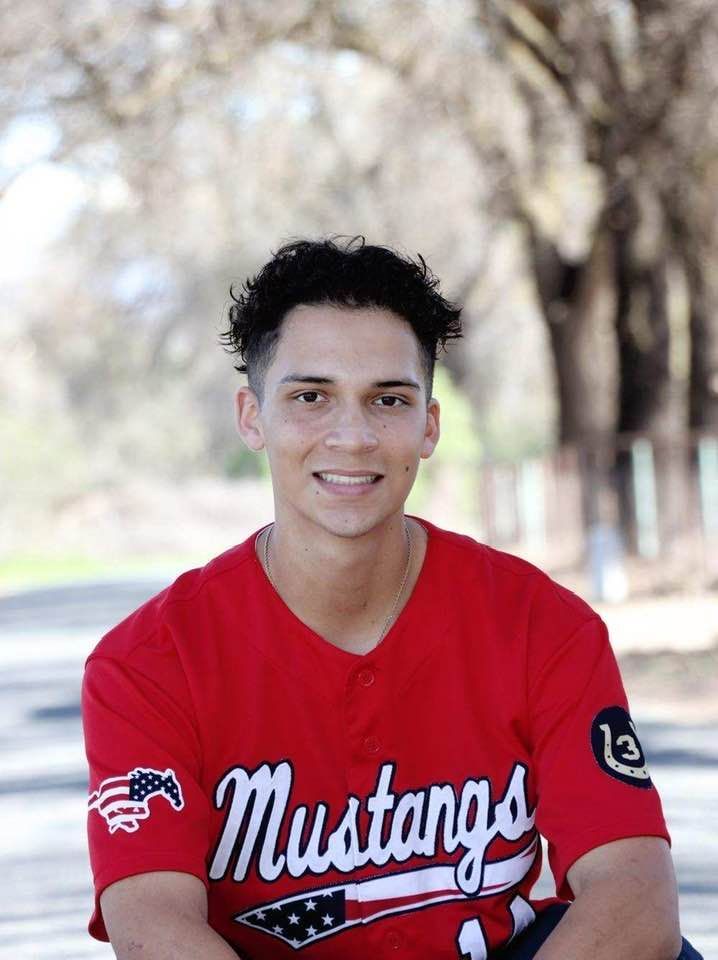 A young man wearing a red mustangs baseball jersey is sitting on a bench.