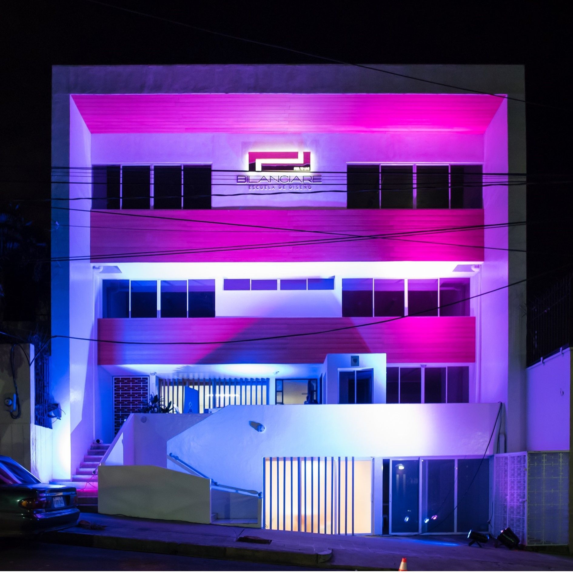Un edificio de varias plantas por la noche, iluminado por luces de acento de color rosa y azul vibrantes, con un logotipo central en la fachada.
