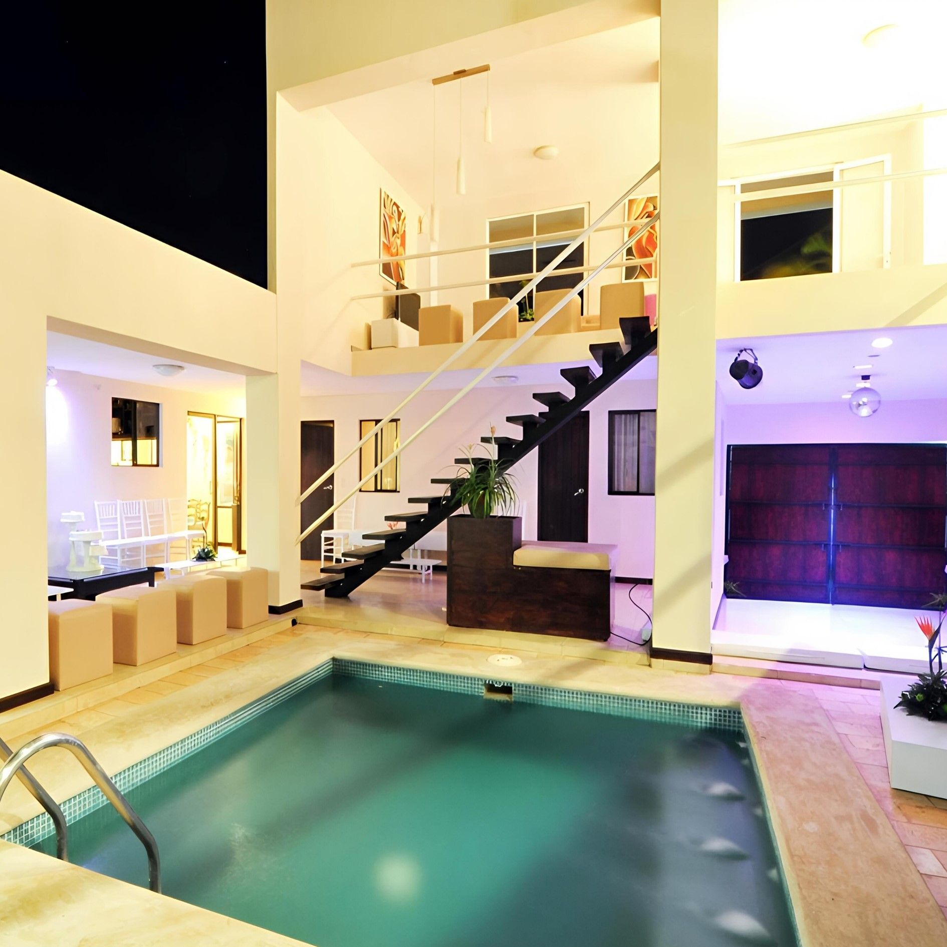 Una piscina cubierta por la noche, junto a una casa moderna con escalera, zona de estar en el patio e iluminación ambiental de color púrpura.