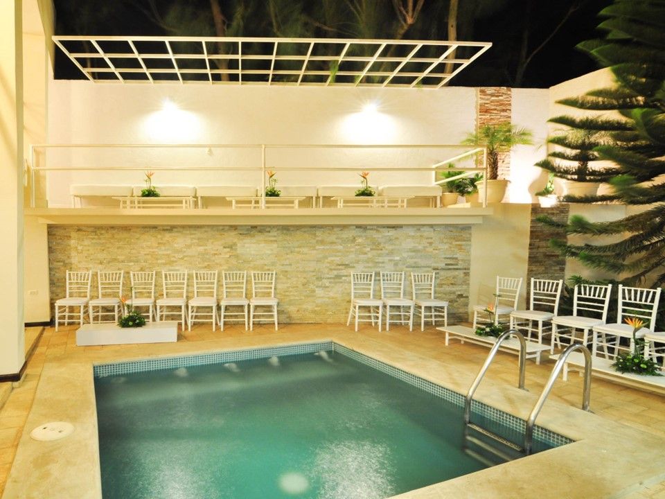 Una piscina al aire libre por la noche, enmarcada por filas de sillas blancas vacías, una pared decorativa de piedra y una pérgola metálica en el techo.