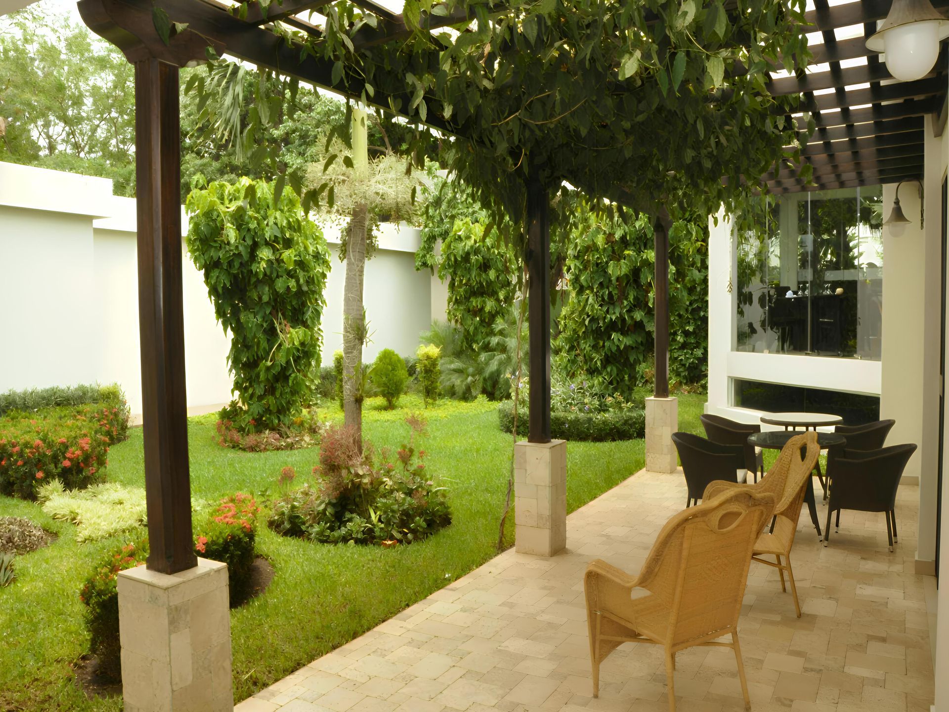 Un patio con suelo de piedra y sillas de mimbre bajo una pérgola, con vistas a un exuberante jardín verde con árboles y arbustos.