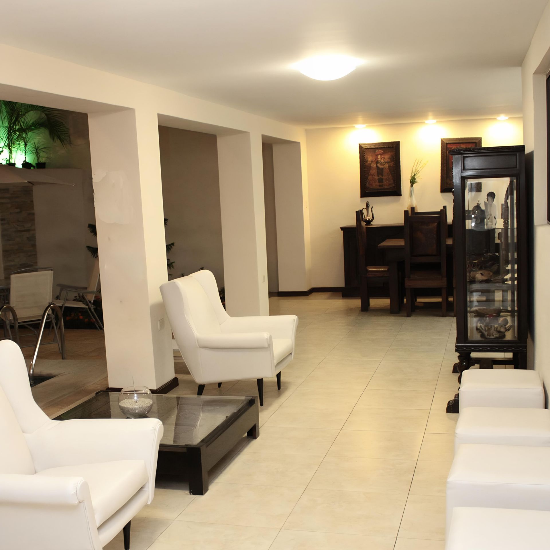 Una sala de estar y comedor con sillones blancos, una mesa de centro baja y una vitrina sobre un suelo de baldosas claras.