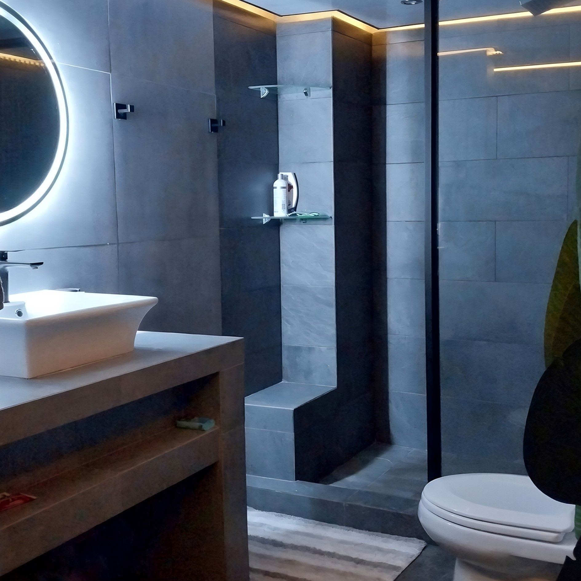 Un baño moderno con iluminación tenue, que cuenta con una ducha de azulejos oscuros, un lavabo sobre encimera de piedra y un espejo redondo retroiluminado.