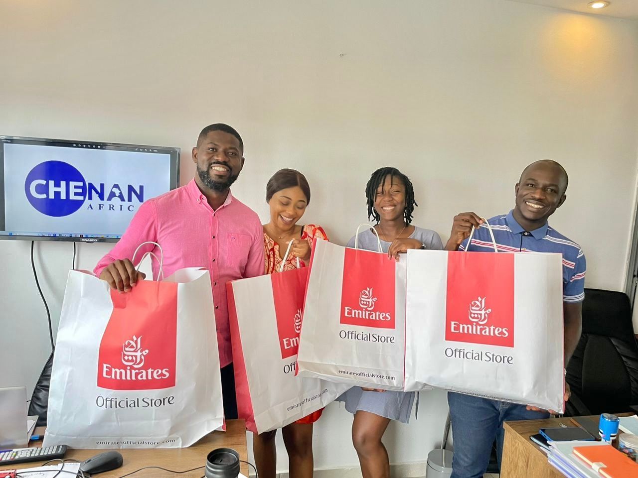 Quatre personnes tenant des sacs de shopping Emirates dans un bureau ; un écran de télévision affiche « MAGASIN DE CHENAN »
