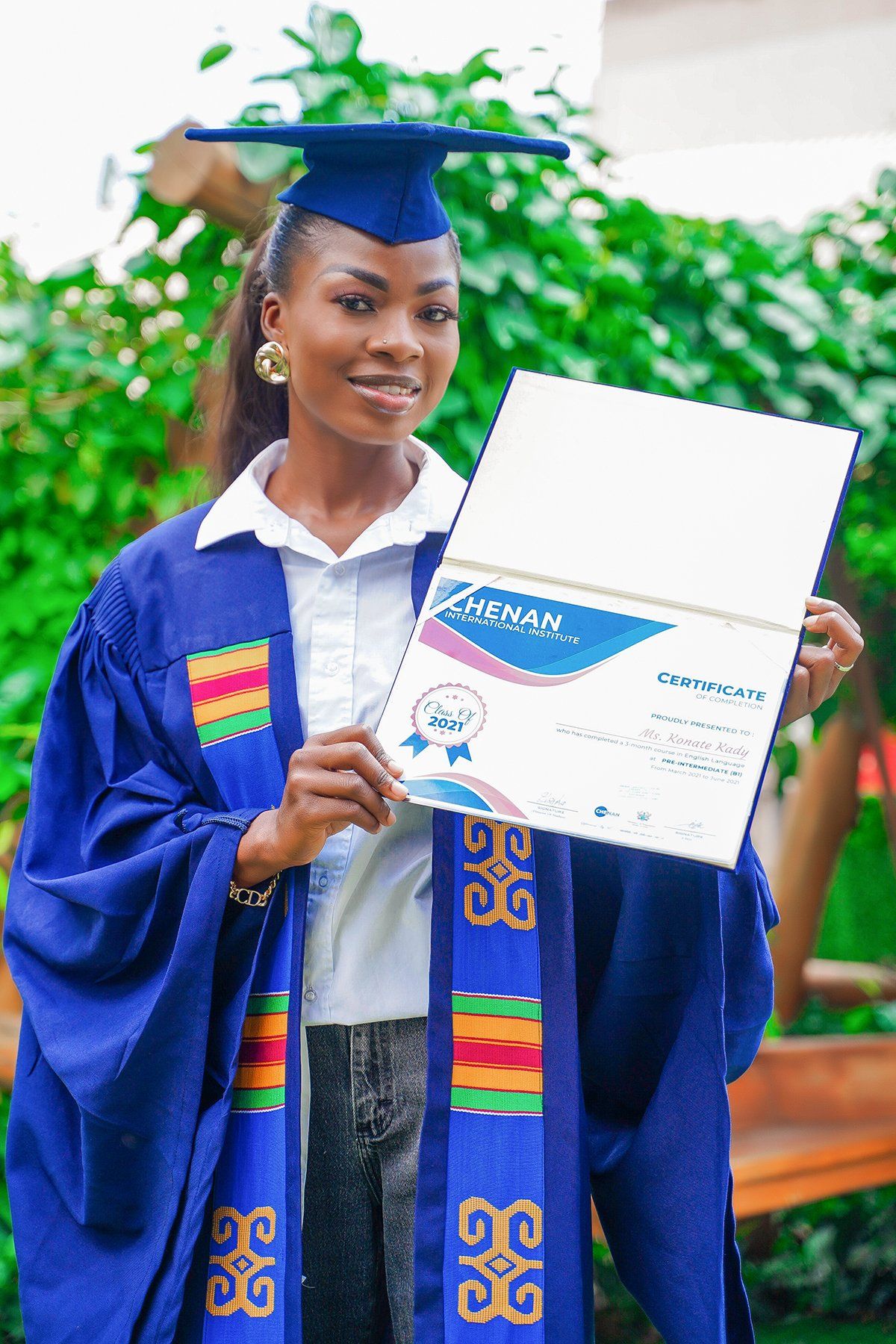 Femme en robe de remise de diplôme, tenant son diplôme, souriante, en extérieur.