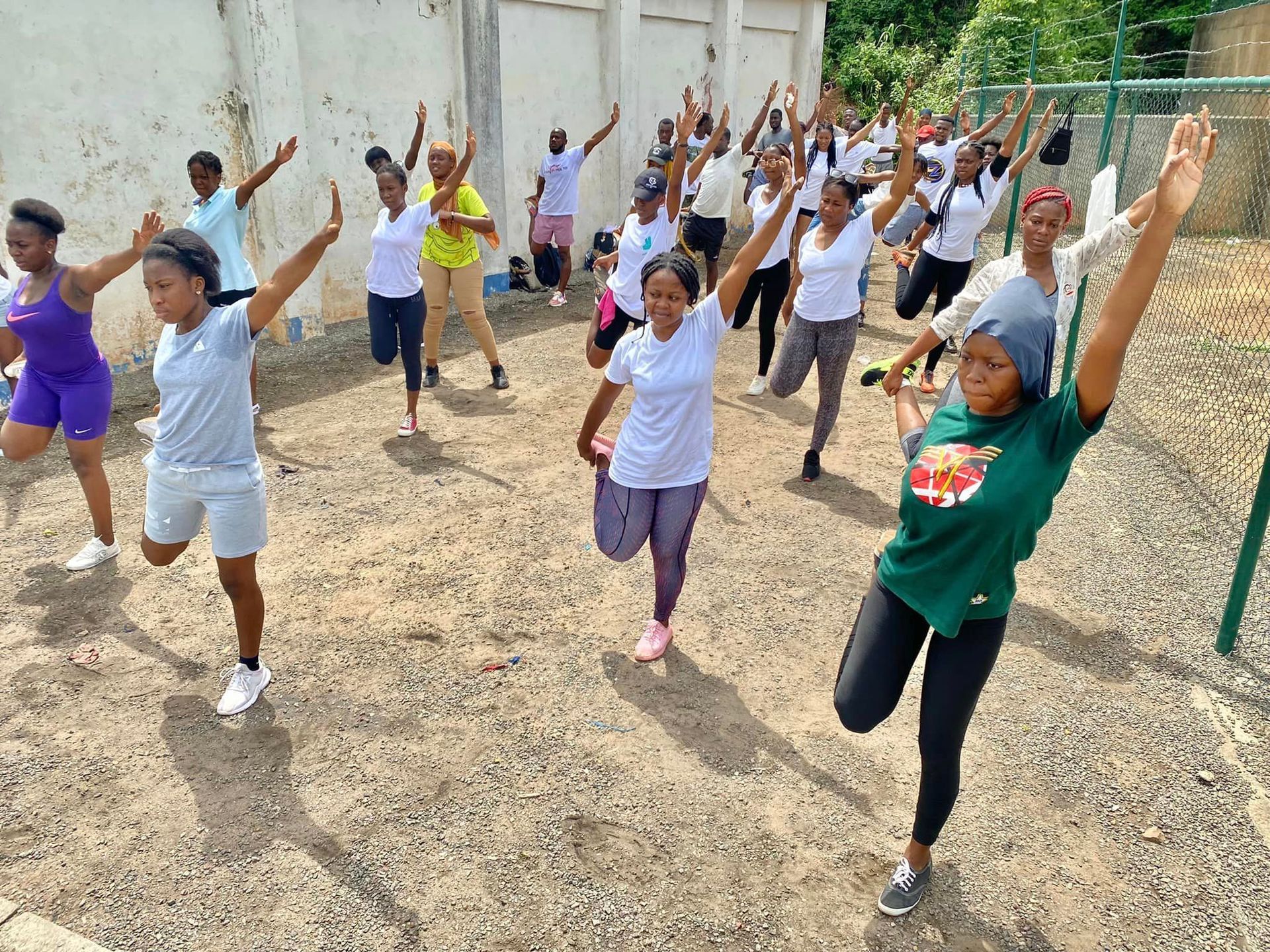 Groupe de personnes faisant de l'exercice en plein air, les bras et les jambes levés. Bâtiment blanc en arrière-plan.