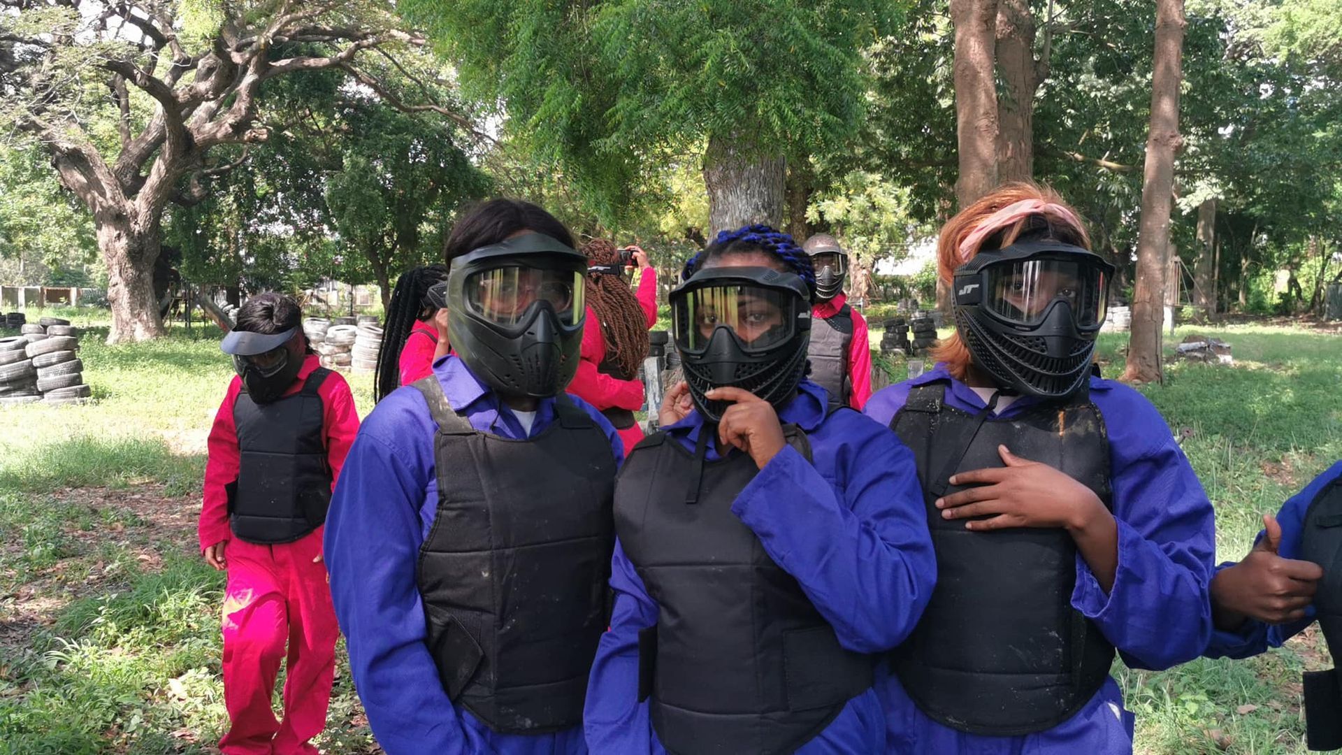 Des personnes en tenue de sécurité posent pour une photo sur un terrain de paintball ; elles portent des masques et des gilets, certaines en combinaisons bleues.