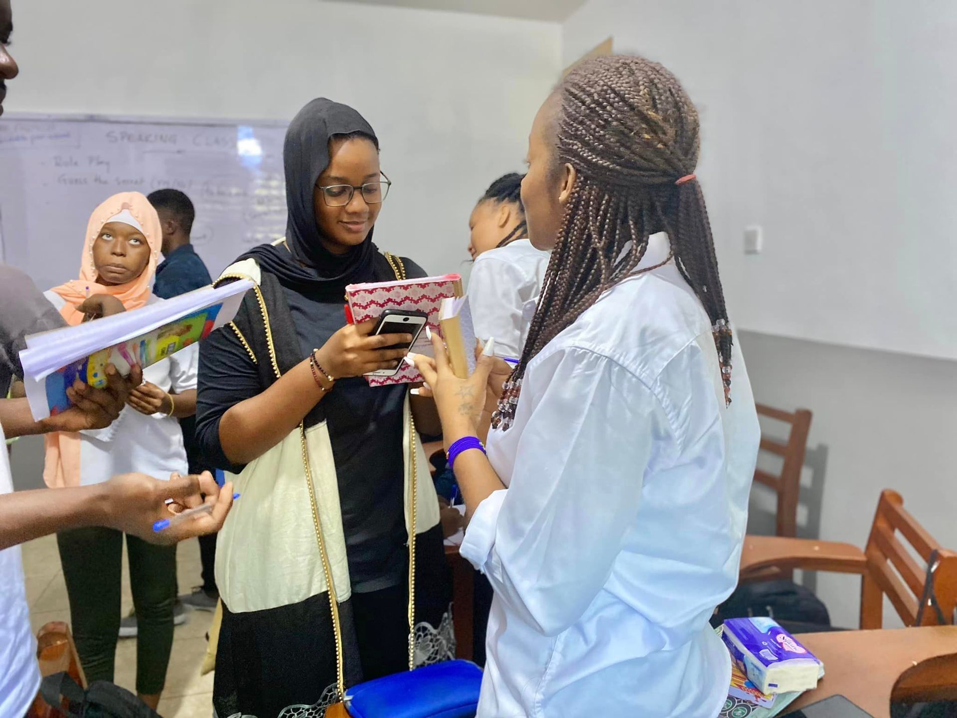 Dans une salle de classe, une femme utilise son téléphone, une autre montre un objet. D'autres personnes les observent.
