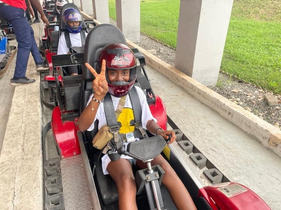 Une personne en karting rouge, casquée, fait le signe de la paix. D'autres personnes sont en file indienne, roulant sur la piste.