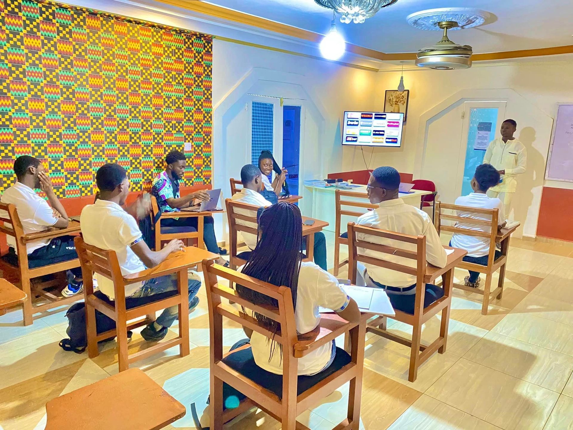 Dans une salle de classe décorée dans un style africain, un groupe de personnes est assis à des bureaux pour une présentation.