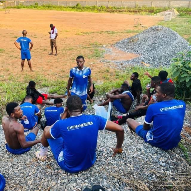 Une équipe de football en uniformes bleus se repose sur un terrain de gravier ; une personne se tient debout à l'arrière-plan.