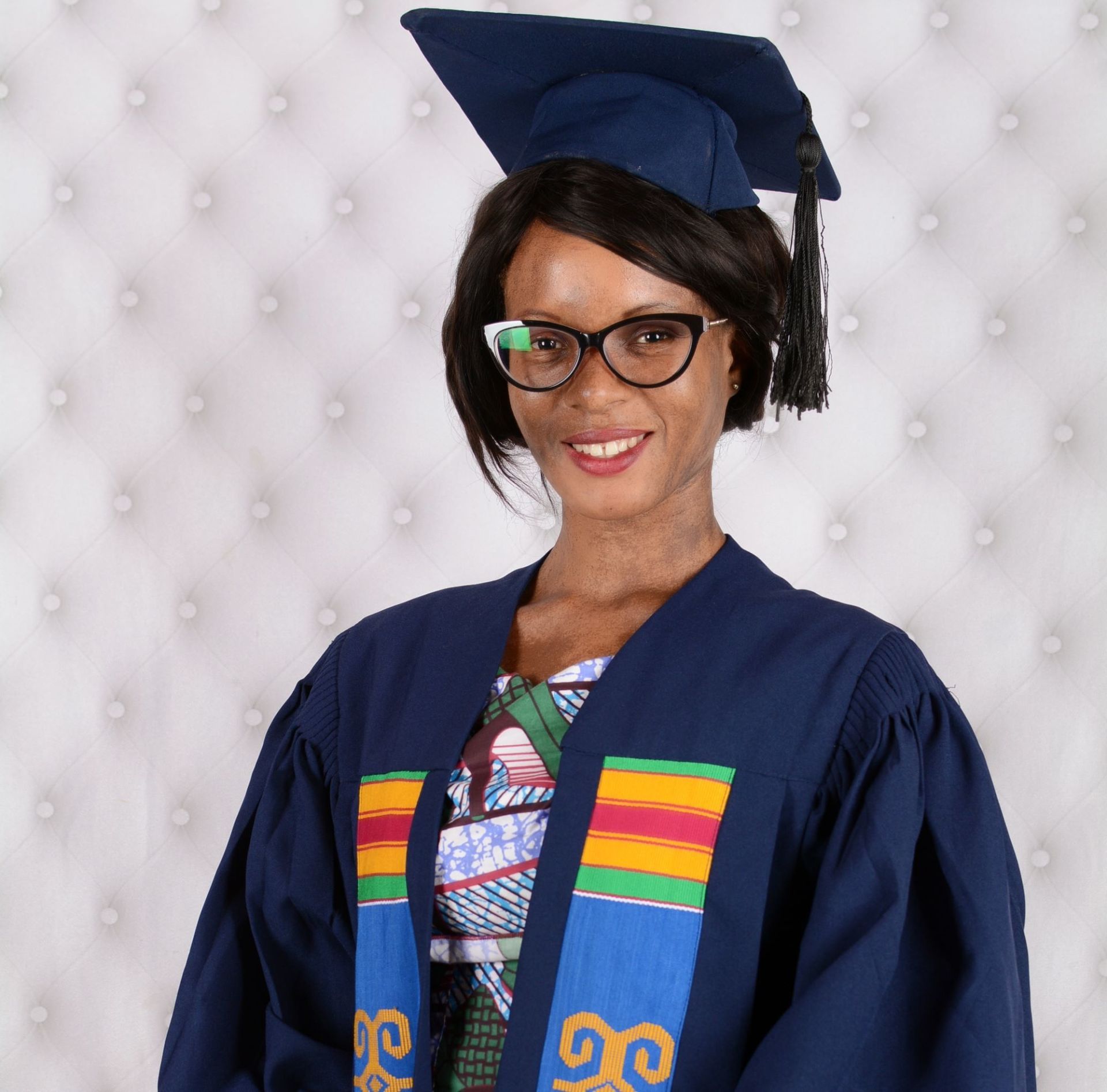 Une femme en tenue de remise de diplôme sourit, portant des lunettes. Elle est coiffée d'une toque et vêtue d'une robe bleu foncé ornée de touches de couleur.