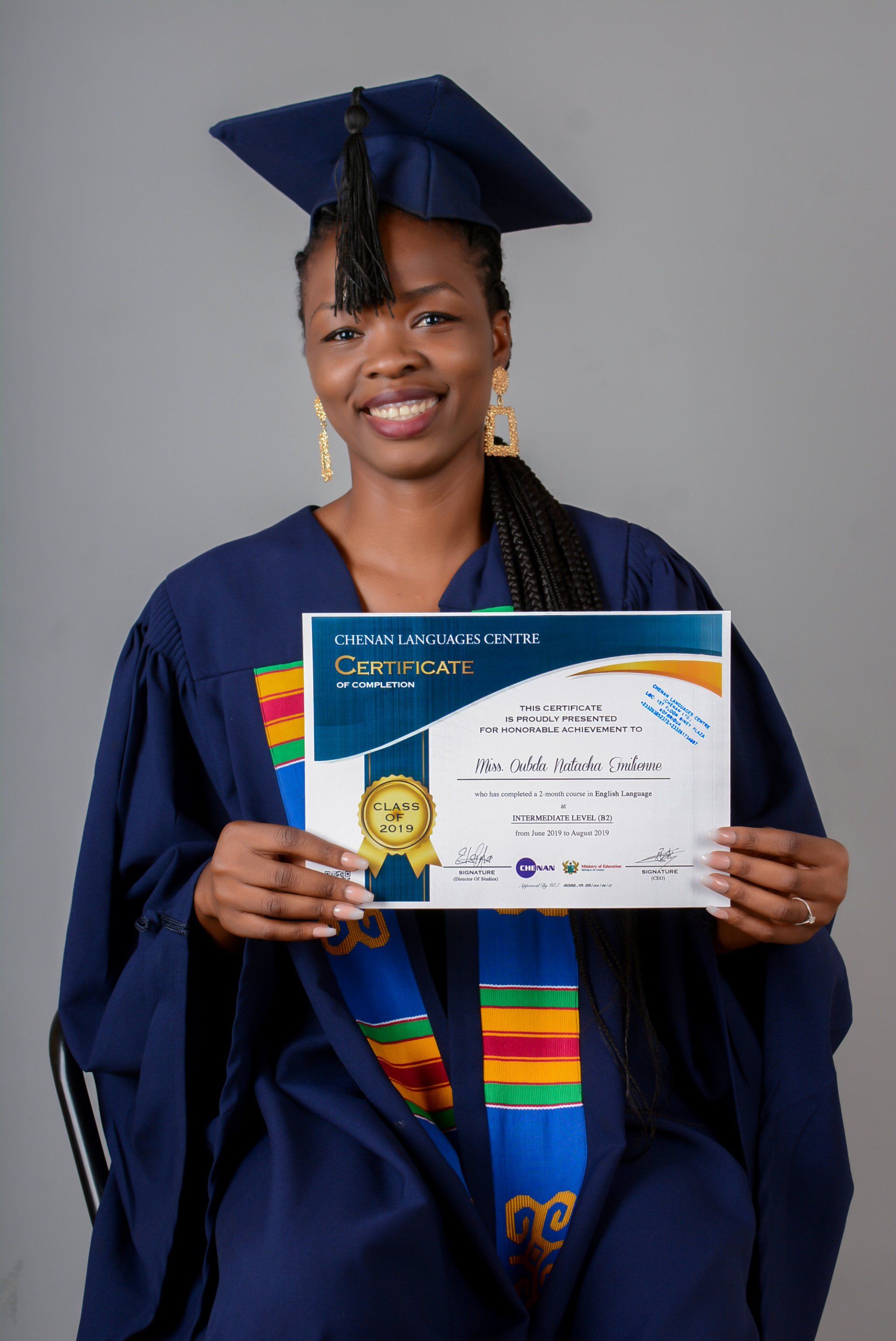 Personne en toge de remise de diplôme, tenant son diplôme et souriante. Fond gris uni.