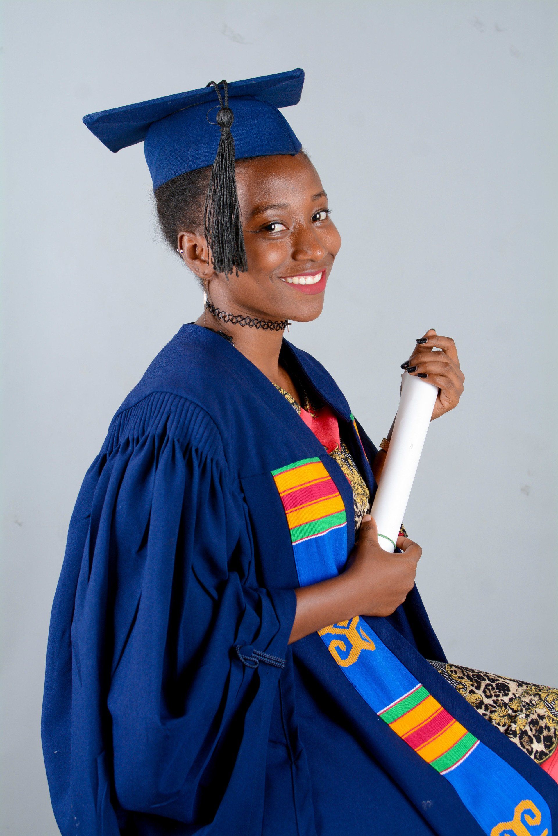 Personne en toge de remise de diplôme, tenant son diplôme et souriante. Robe, toque et écharpe bleu foncé à motifs colorés, sur fond clair.