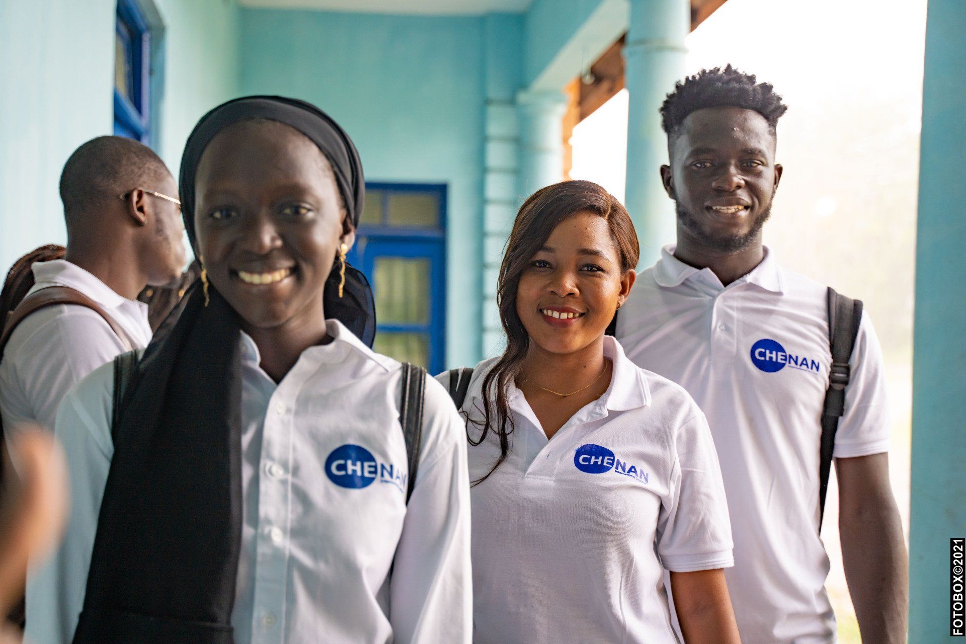 Trois personnes souriantes dans un couloir, vêtues de chemises blanches à logo, regardent l'objectif.
