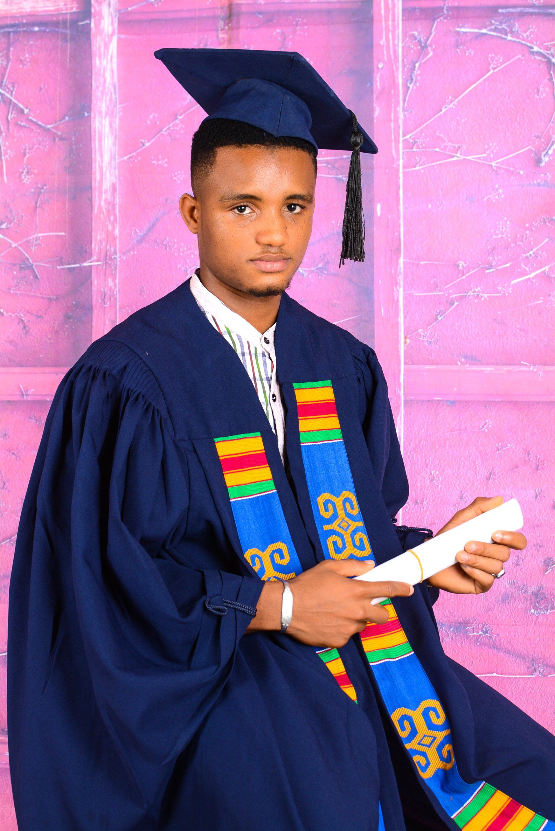 Personne en tenue de remise de diplôme tenant un diplôme : robe bleue, toque noire, écharpe colorée, fond rose.