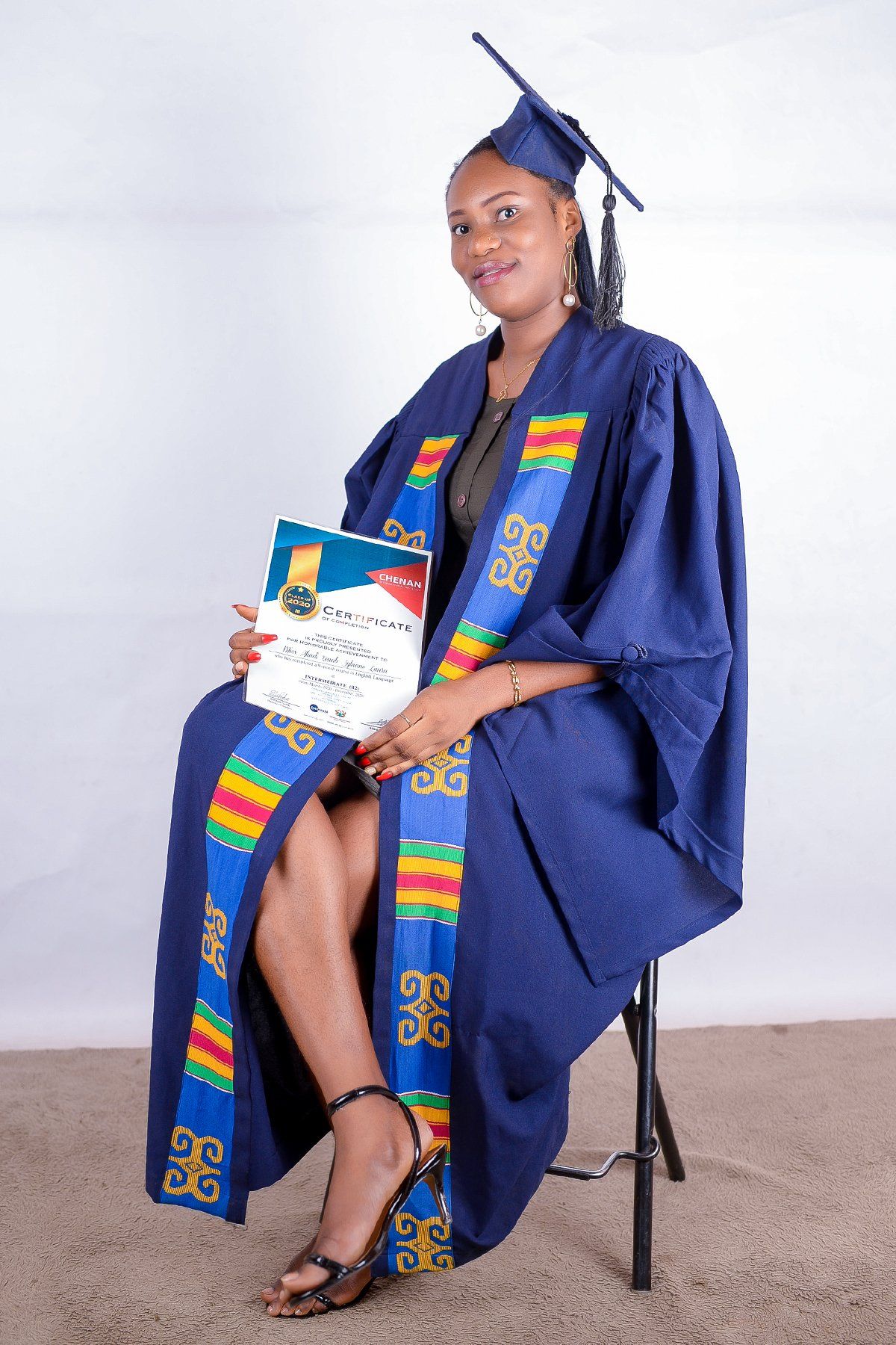 Personne en toge de remise de diplôme, tenant un diplôme, assise devant un fond blanc.