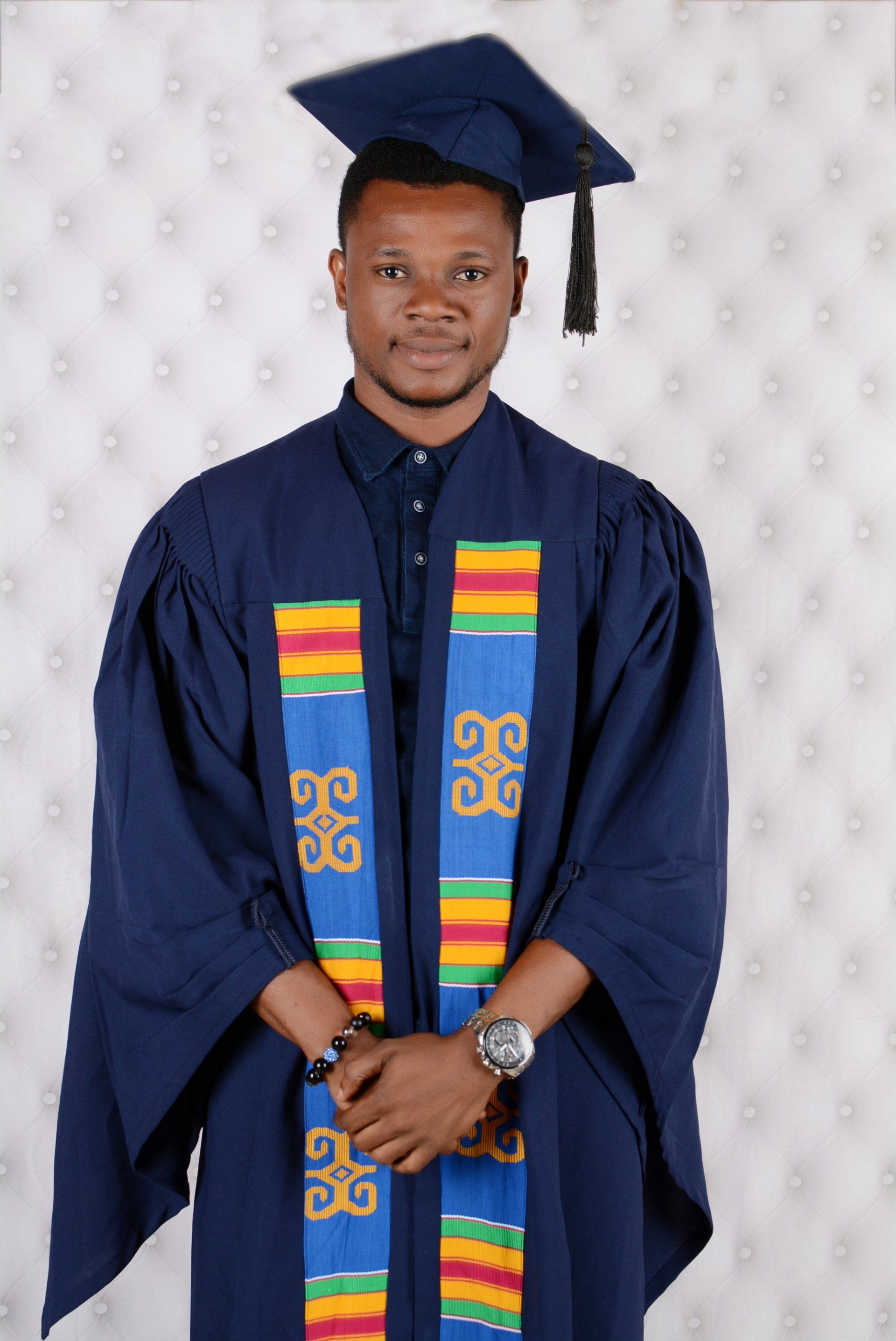 Homme en toge et mortier de remise de diplôme, debout, souriant. Écharpe bleue et colorée, montre-bracelet.