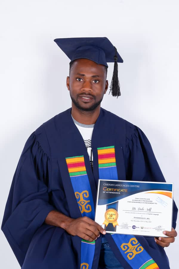 Homme en toge et chapeau de diplômé tenant un certificat, souriant. Fond blanc.
