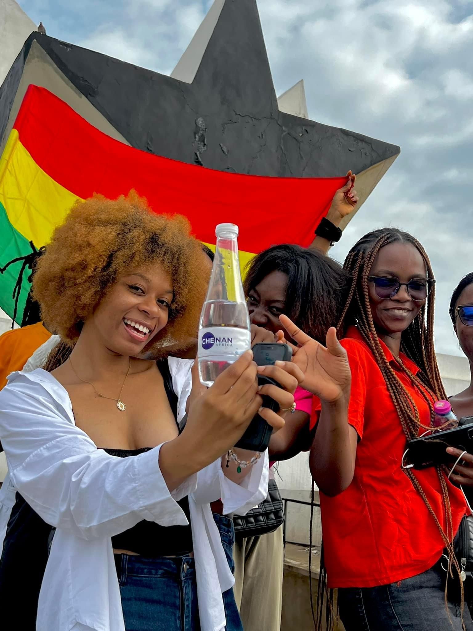 Un groupe de personnes prend un selfie avec le drapeau ghanéen en arrière-plan. Une femme tient une bouteille d'eau.