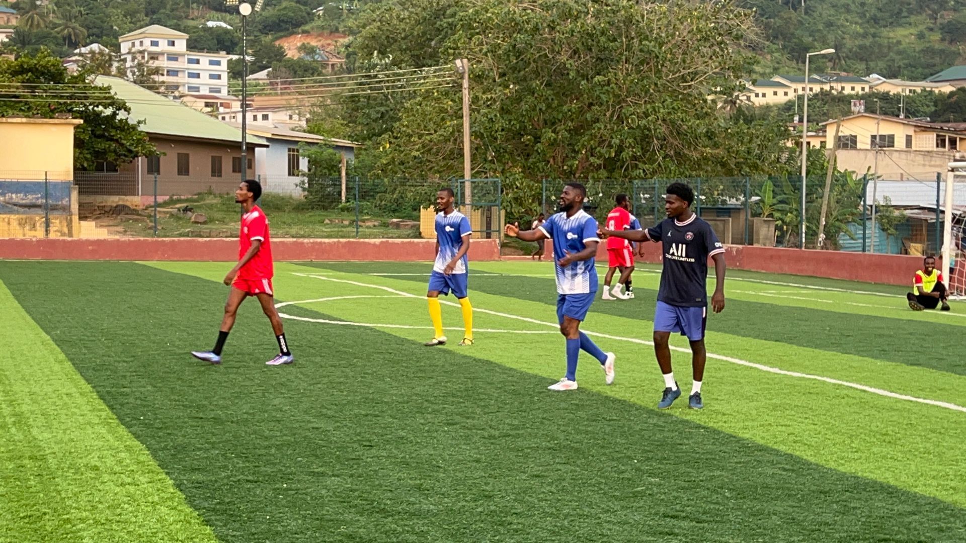 Des joueurs de football évoluent sur un terrain en gazon synthétique vert. Ils portent des maillots bleus et rouges, marchent et s'arrêtent. Des maisons se dressent à l'arrière-plan.