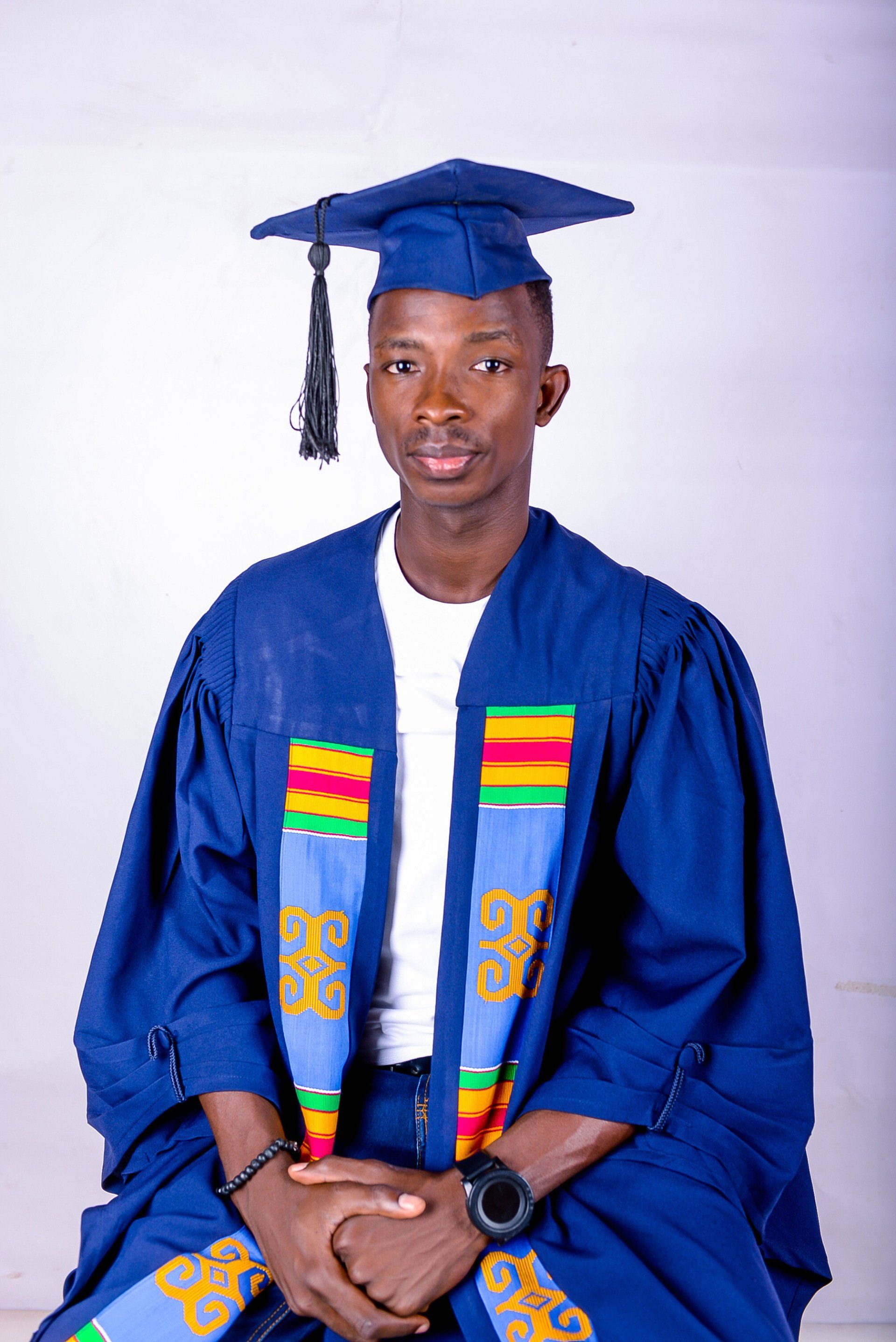 Un homme portant une casquette et une robe de graduation est assis, les mains jointes.