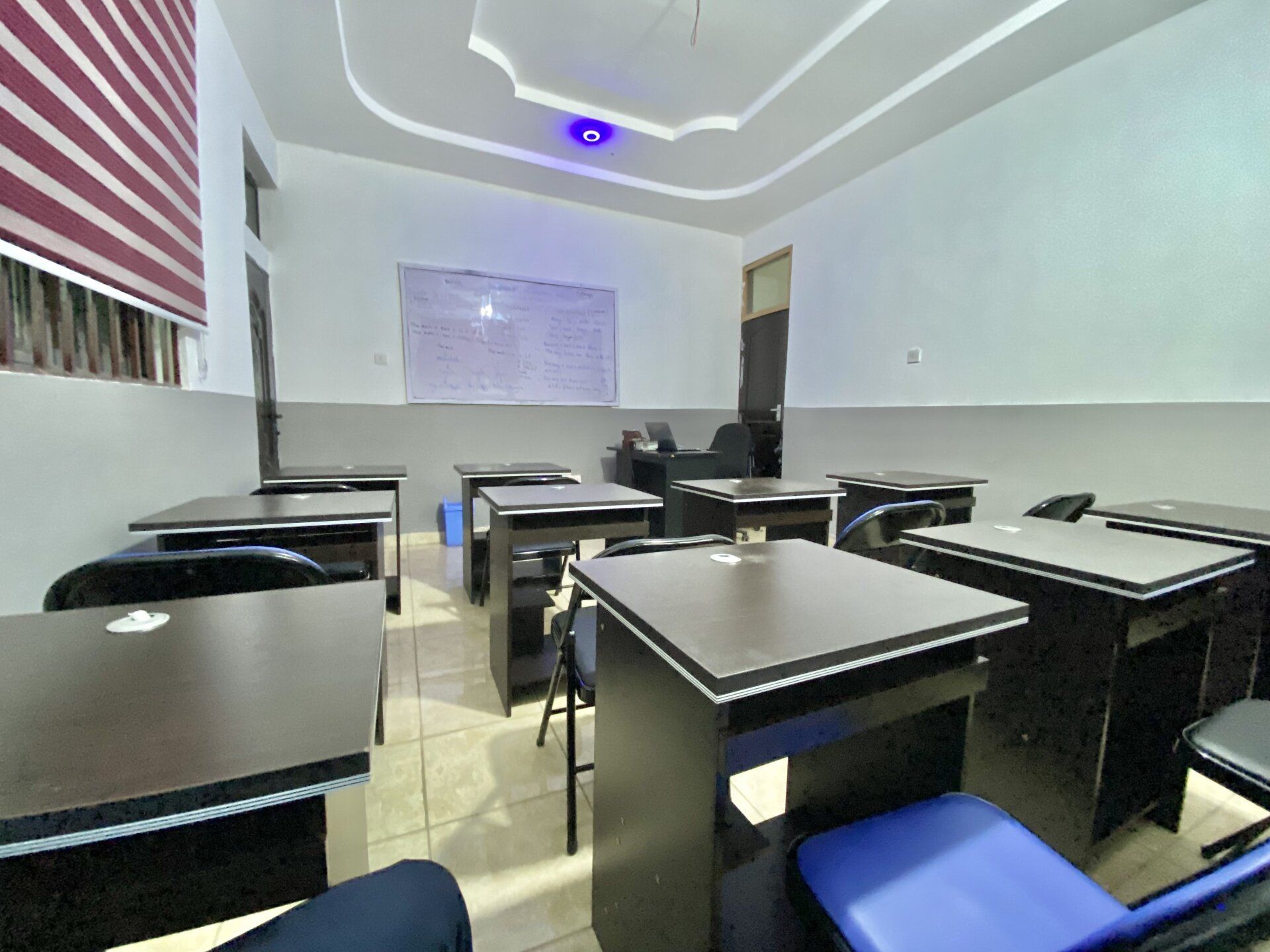 Salle de classe avec des rangées de tables et de chaises. Tableau blanc au mur, stores à la fenêtre.