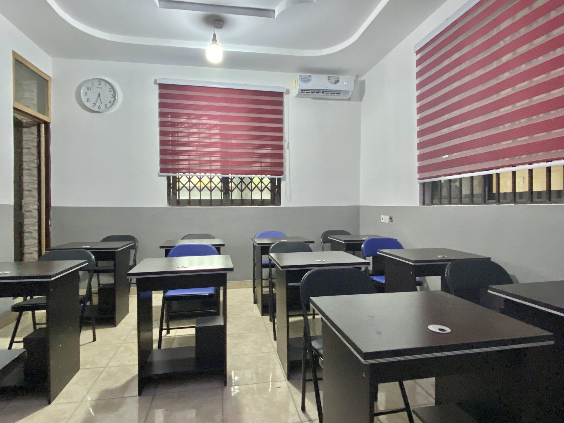 Salle de classe vide avec des rangées de pupitres, des fenêtres à stores et une horloge au mur.