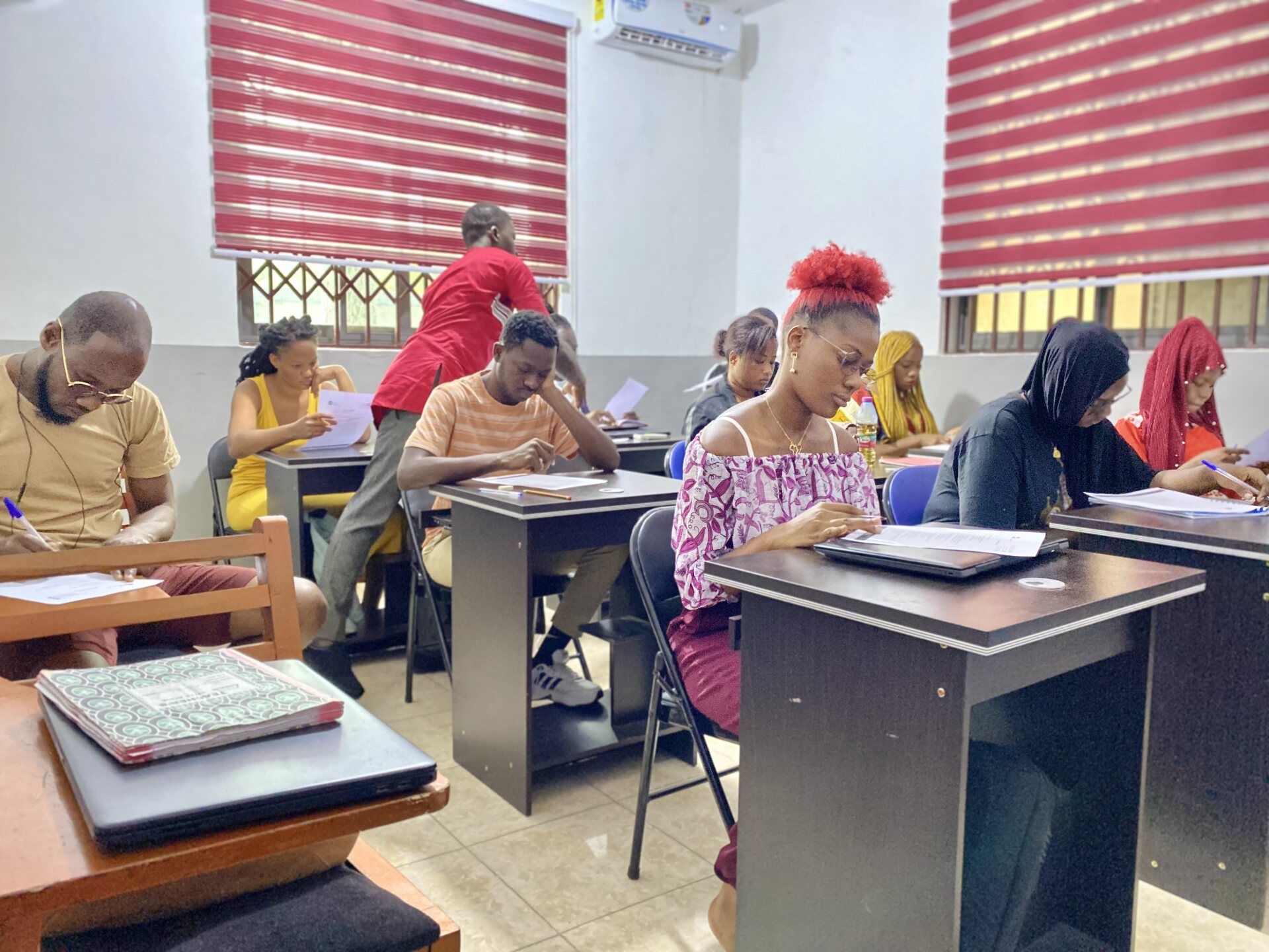 Des élèves sont assis à leurs pupitres dans une salle de classe, certains écrivent. Un professeur passe près d'un élève.
