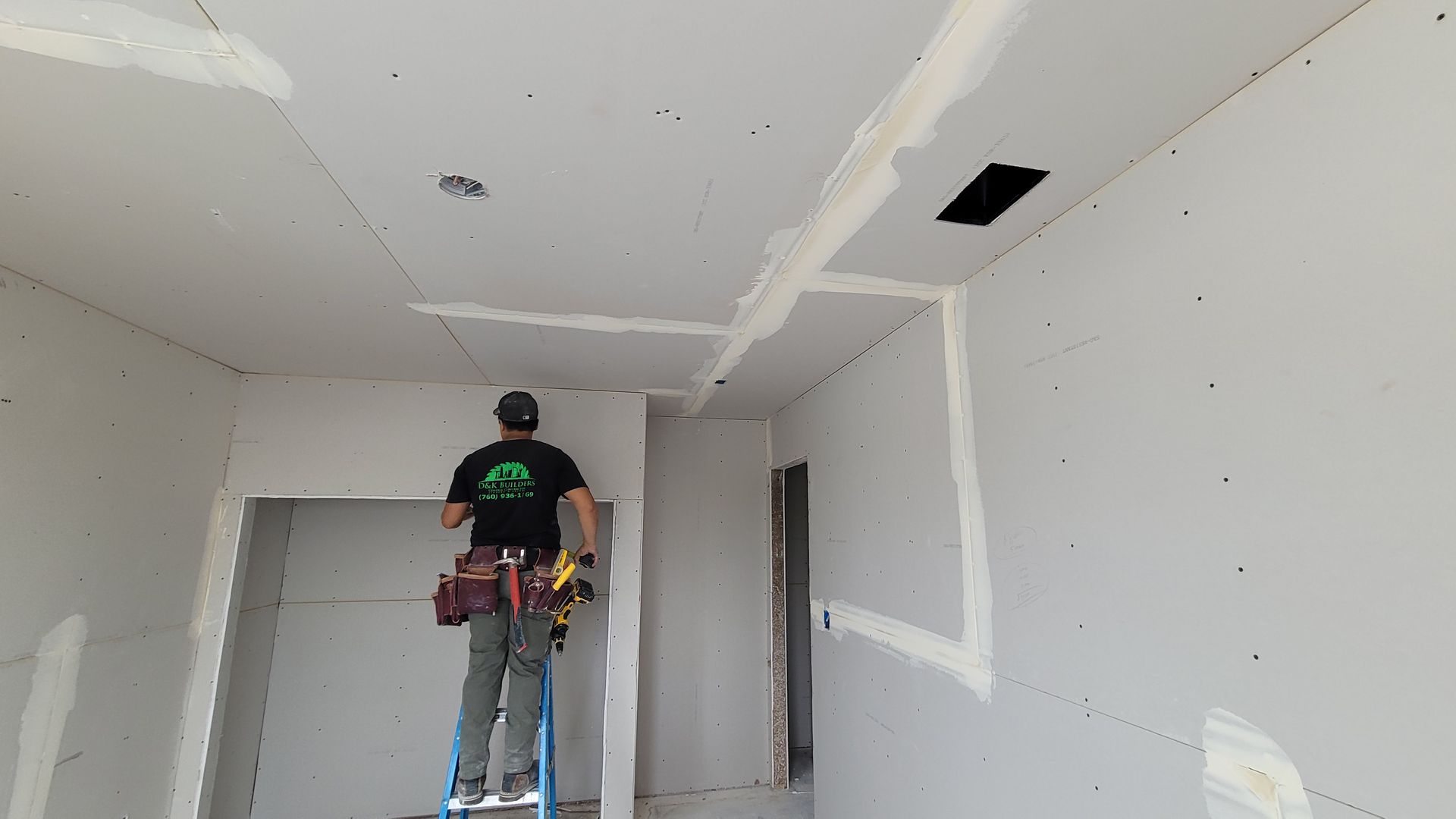 A worker on a ladder installs drywall on the ceiling and walls of an unfinished room with visible taped seams.