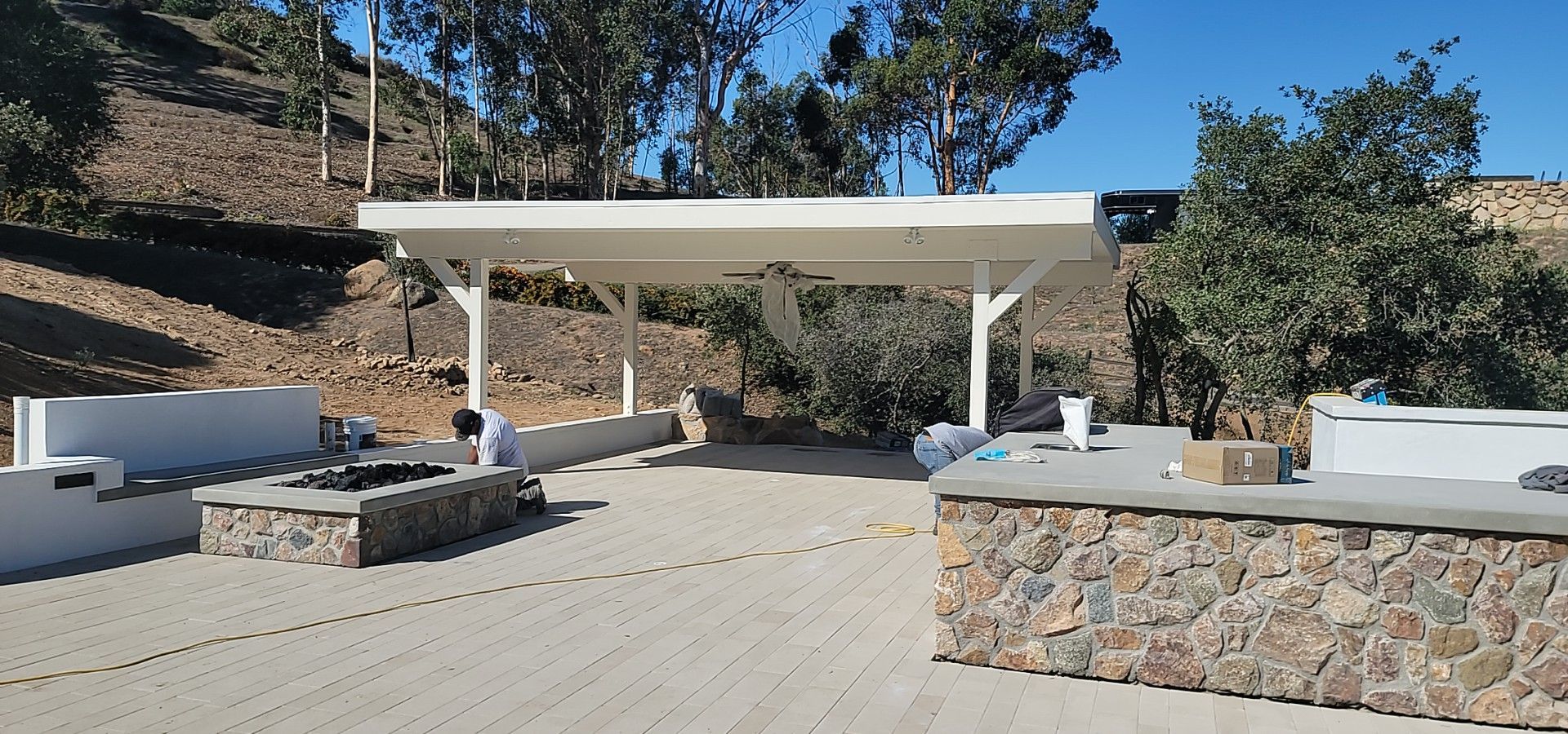 A white open-air patio structure with stone base counters and gravel flooring sits in a hillside landscape.