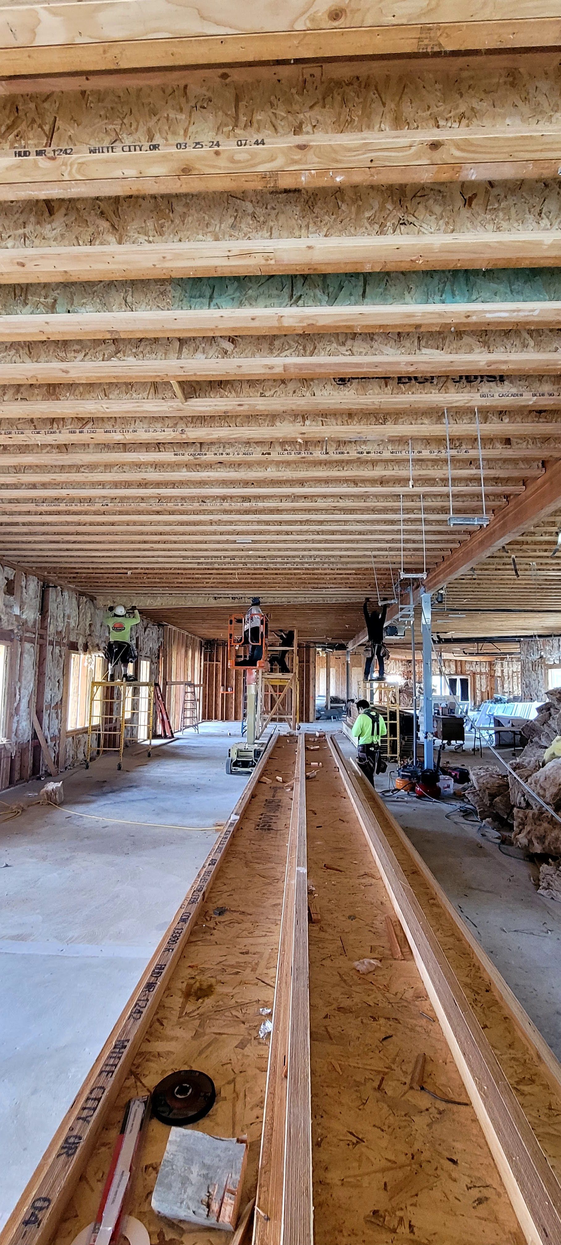 A view down a long, central wooden beam inside an unfinished building construction site with exposed joist ceilings.