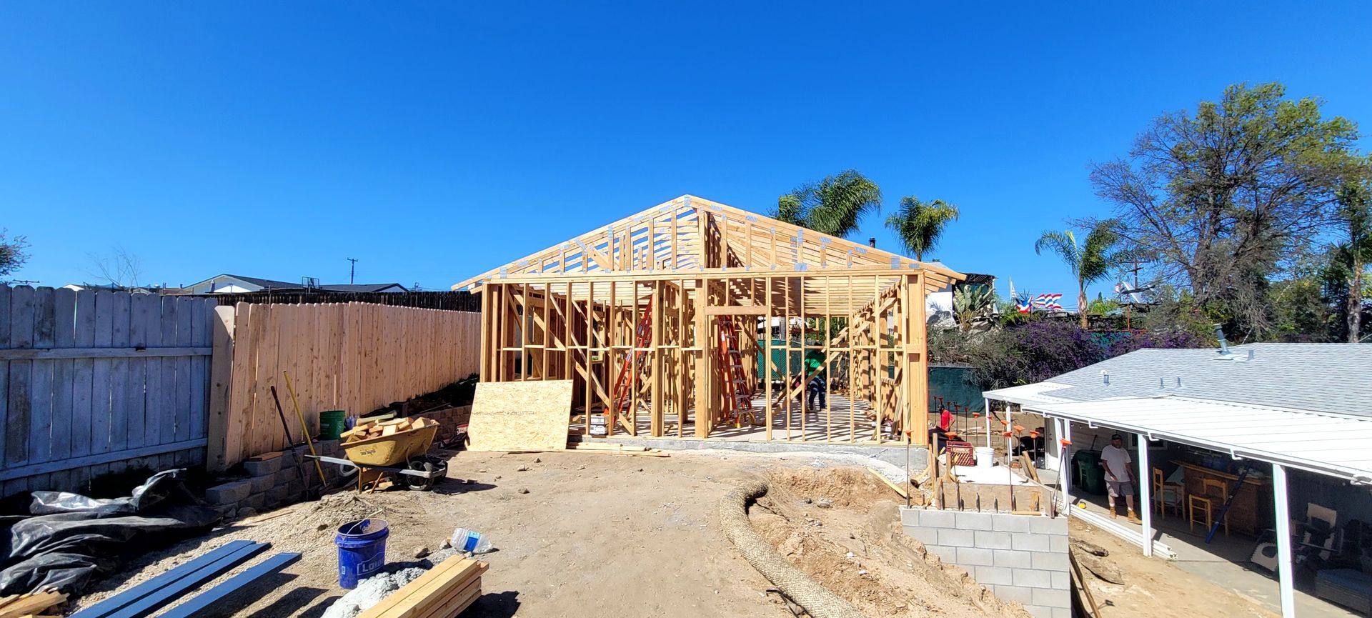 A house under construction with exposed wooden framing stands in a yard under a clear blue sky.