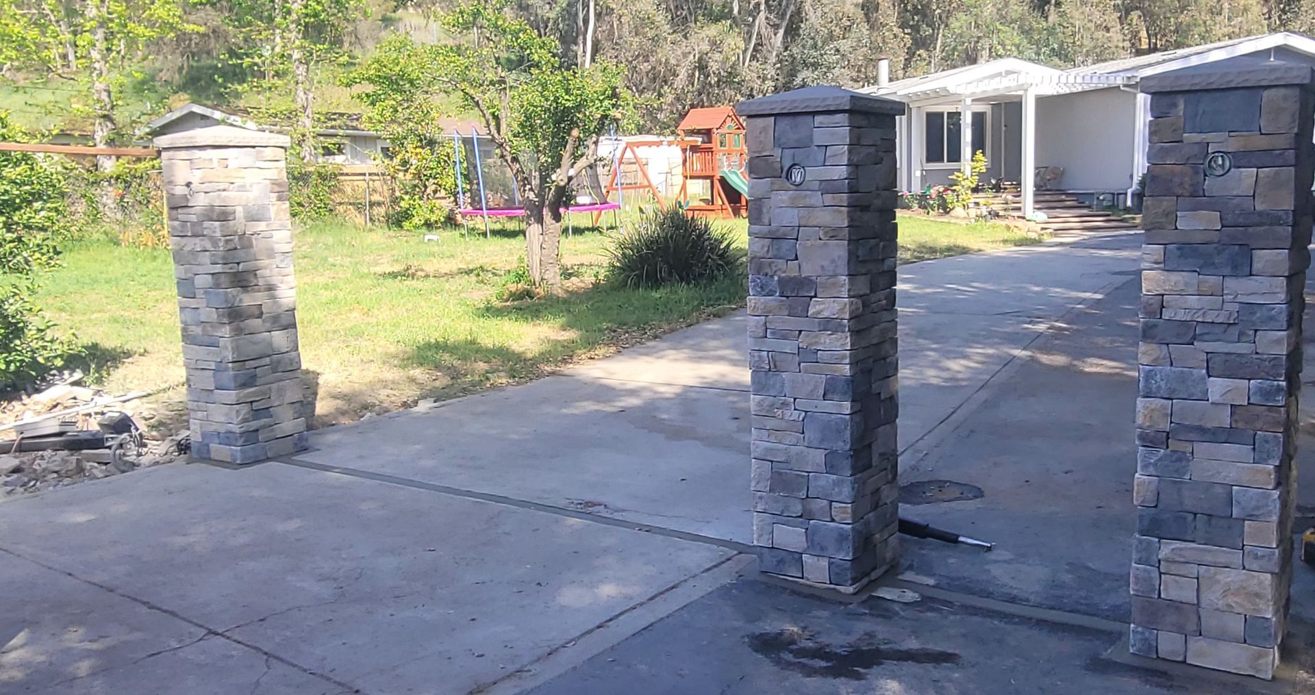 Three stone pillars stand at the entrance of a concrete driveway leading to a house with a green lawn and trees.