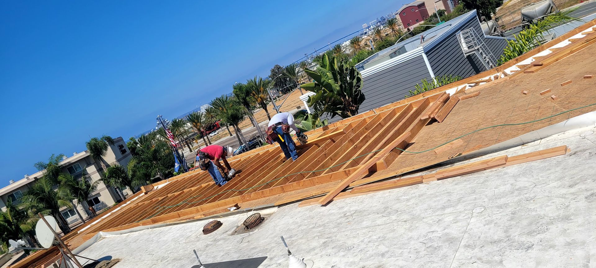 Three people work on a sloped wooden frame construction site near palm trees and buildings under a clear blue sky.