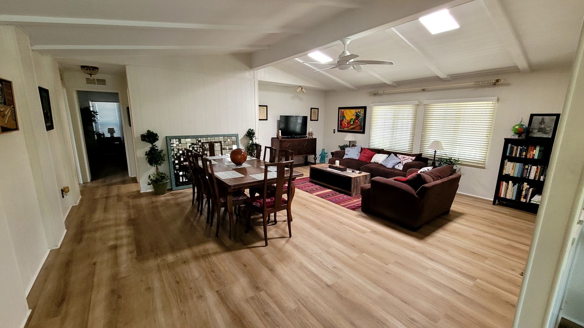 An open-concept living and dining room with wood-look flooring, a dining table, a dark sofa, and a bookshelf.