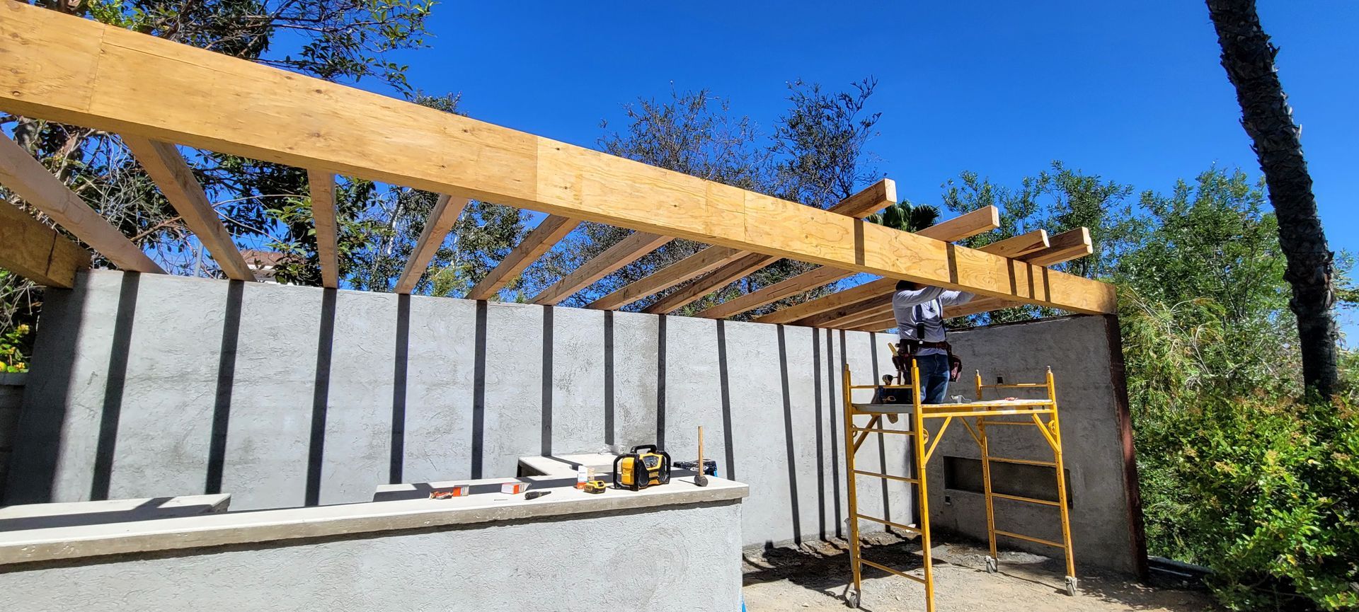 A worker stands on yellow scaffolding under a partially constructed wooden roof frame against a gray stucco wall.