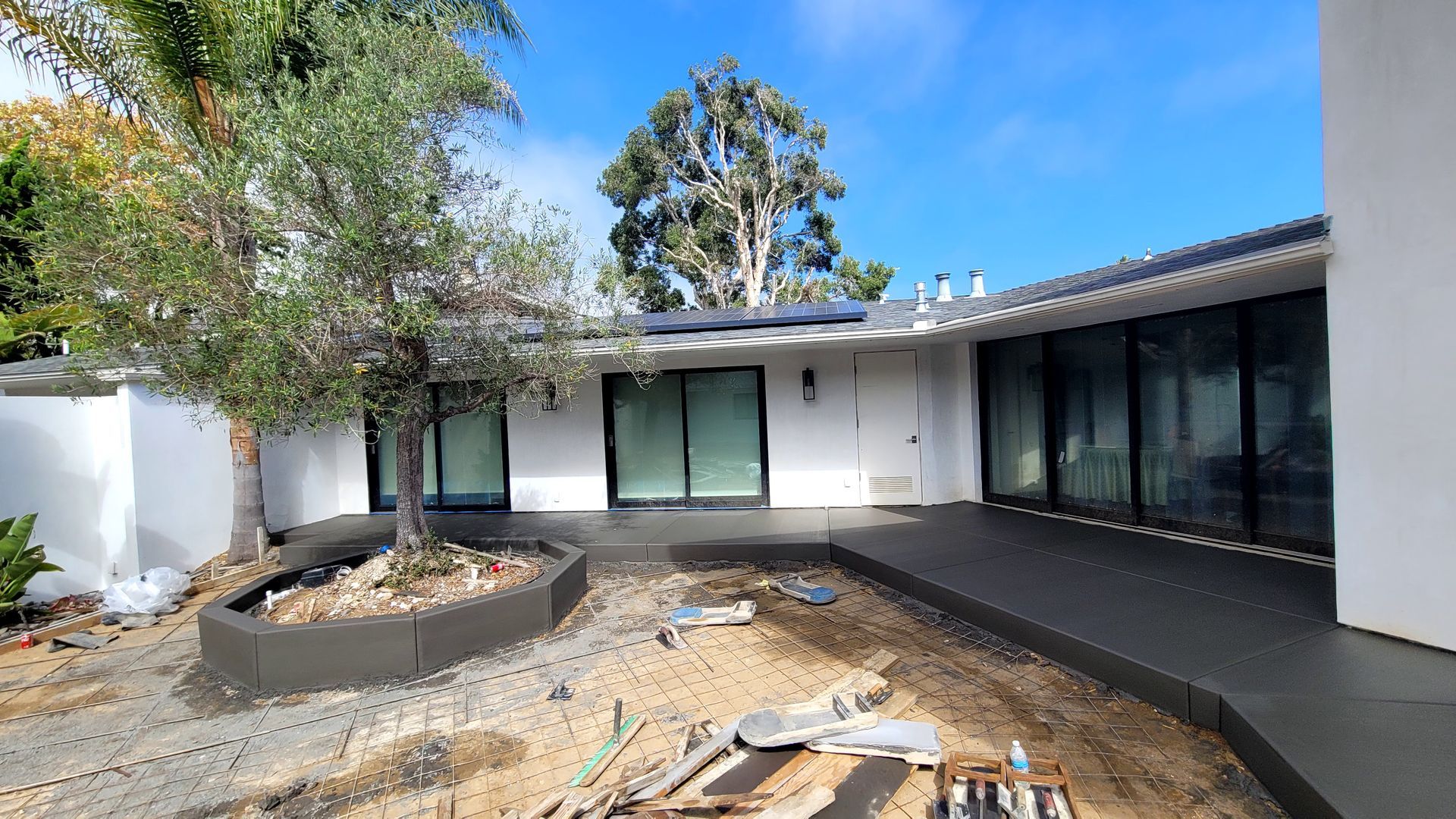 A white house exterior featuring a newly poured dark concrete patio, a central tree planter, and black-framed glass doors.