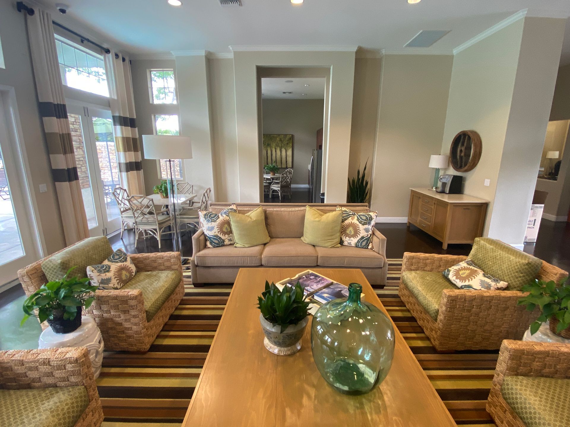A cozy, sunlit living area with a tan sofa, woven armchairs, a wooden coffee table, and a large decorative glass vase.