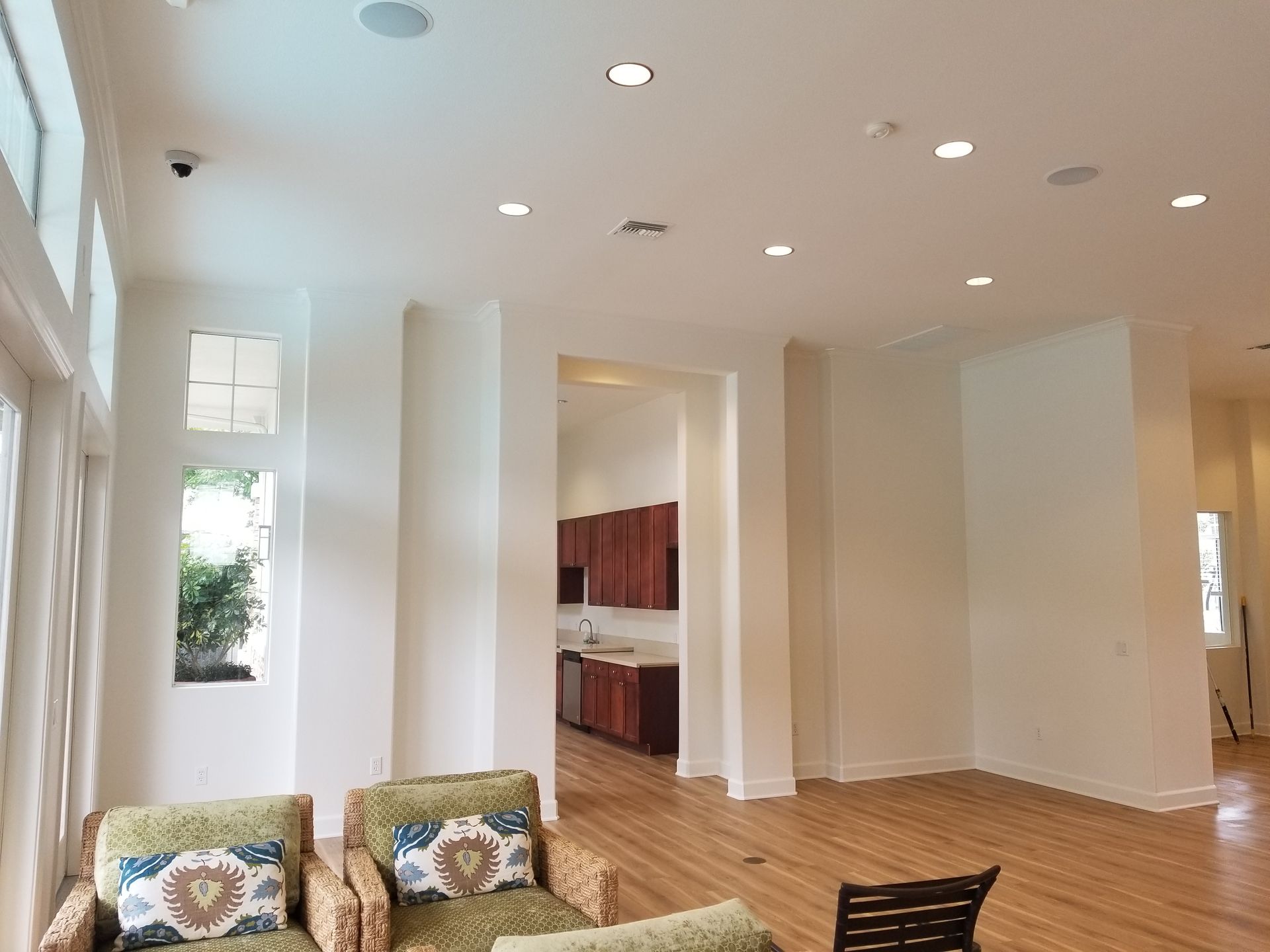 A bright, spacious room with white walls, light wood floors, recessed lighting, and a view into a wooden kitchen area.