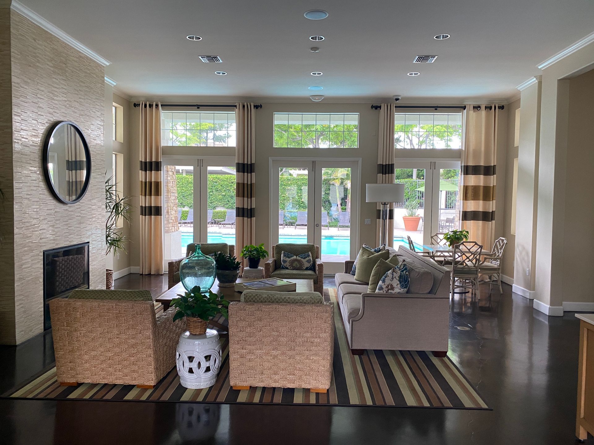 A bright, spacious living room with neutral-toned furniture, a stone fireplace, and floor-to-ceiling windows with pool view.