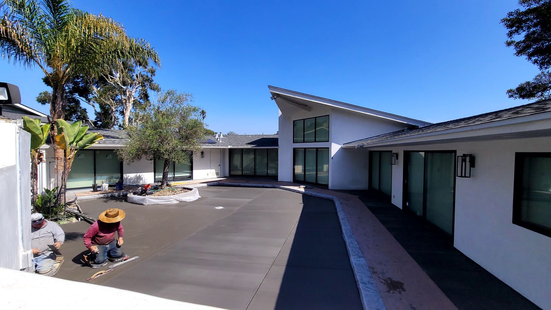 Two workers in hats kneel on a dark concrete driveway in front of a white, modern single-story home under a clear sky.