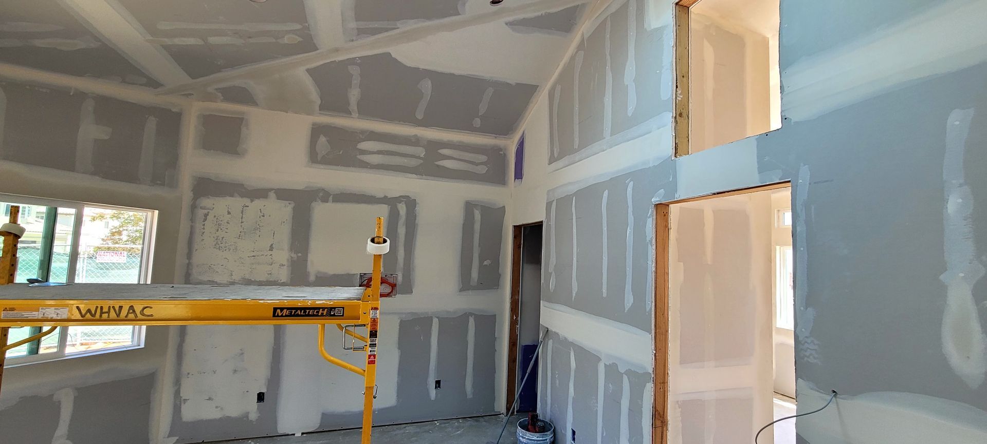 An interior room under construction with newly installed grey drywall panels and a yellow scaffolding tower.