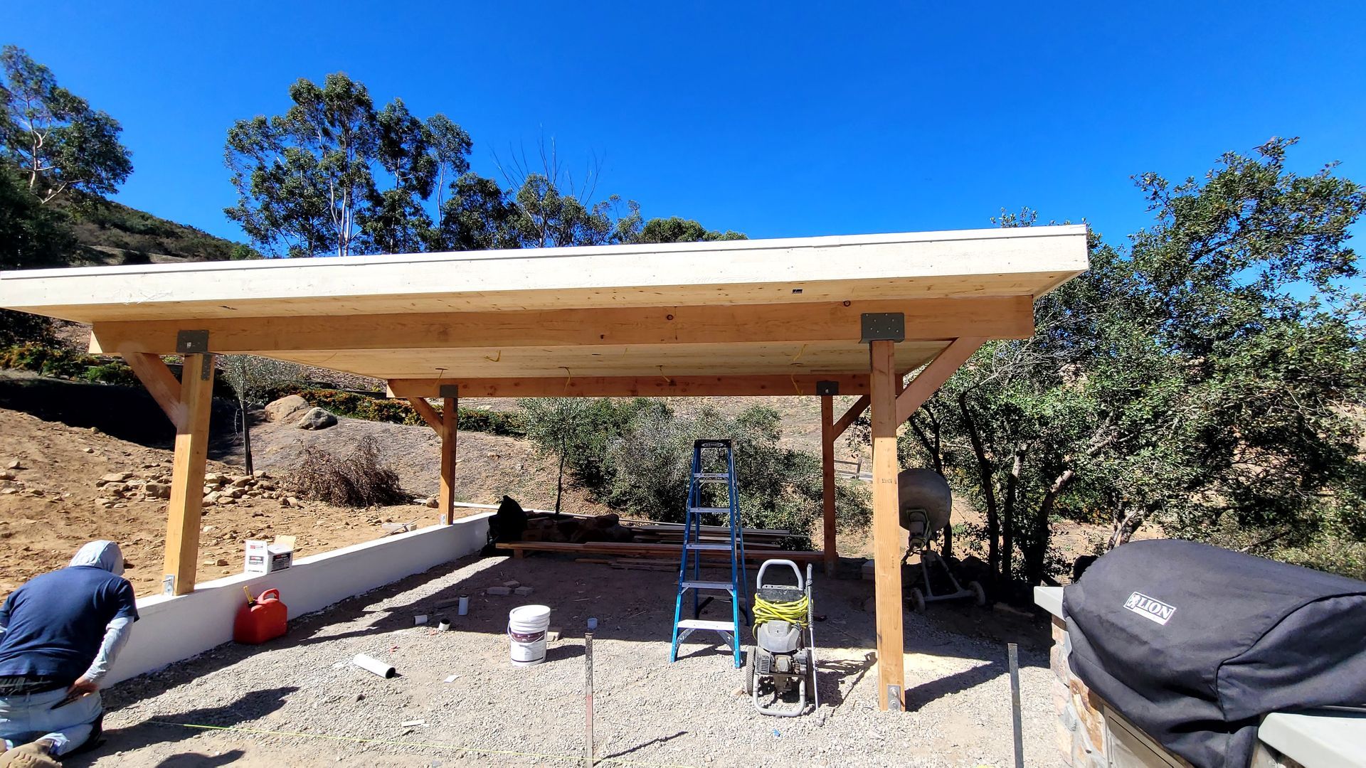 A wooden patio cover frame being built over a gravel area next to a person, a ladder, and a grill under a clear blue sky.