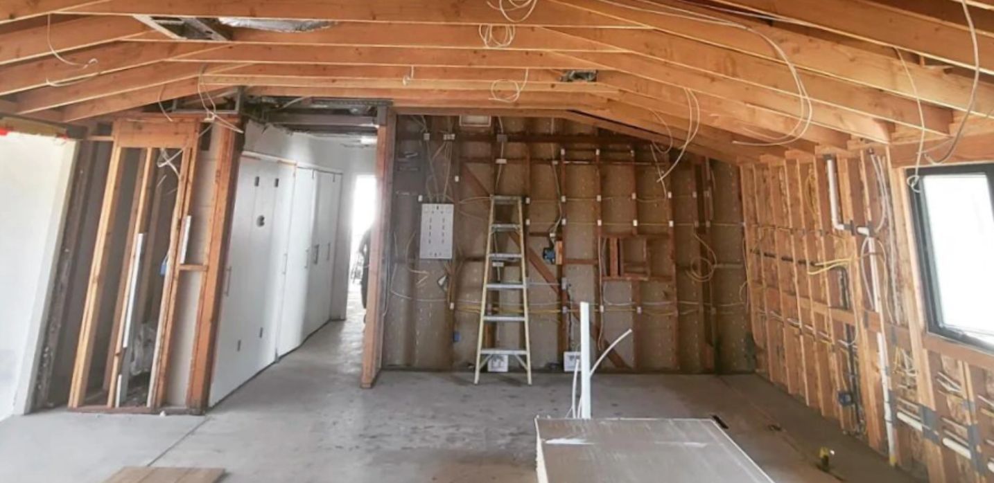 An interior view of a room under construction, showing exposed wooden wall studs, rafters, and a ladder against a wall.