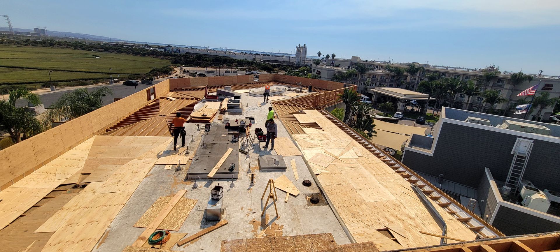 Workers construct the wooden roof deck of a new building under a clear blue sky, overlooking a suburban landscape.