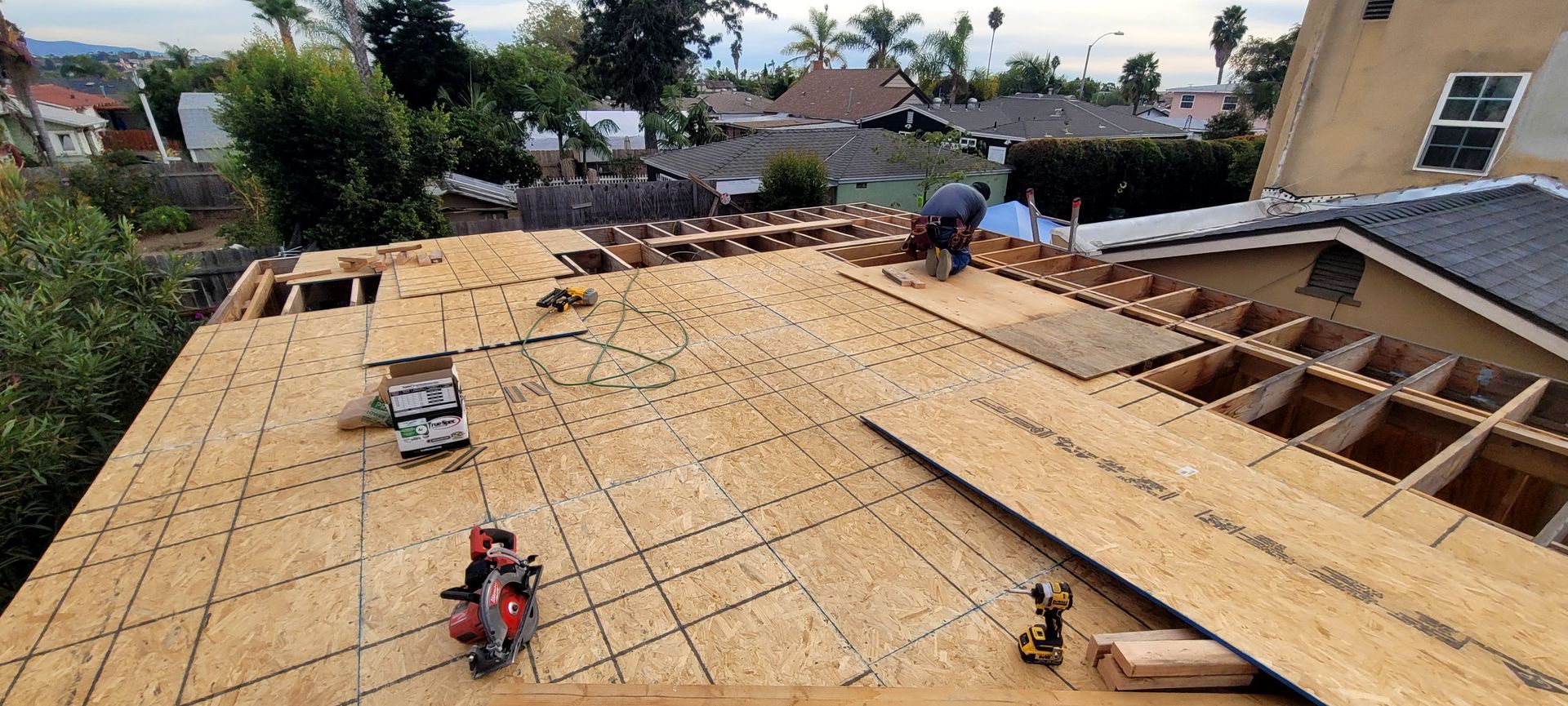 A construction worker installs plywood sheathing onto wooden roof rafters on a sunny day.